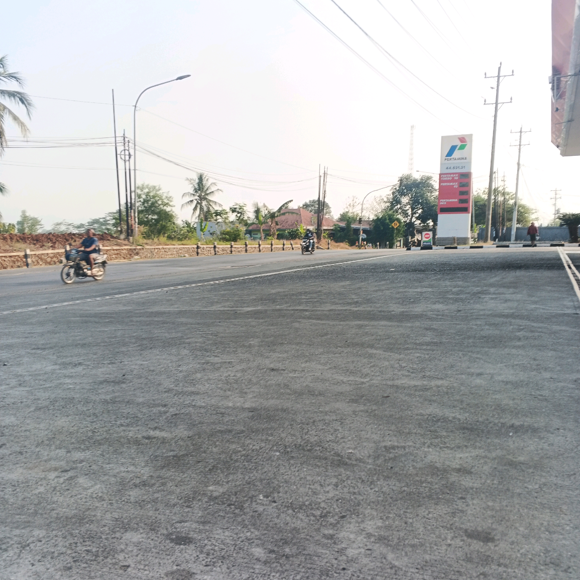 The image depicts a street scene in Kebasen, Indonesia. A man is riding a motorcycle on the asphalt road, wearing a blue shirt. In the background, a Pertamina gas station sign is visible, with prices displayed for different fuel types, including Pertamax Turbo and Pertamax. Several utility poles with electrical wires are also present. The environment includes scattered trees, houses, and a concrete barrier along the roadside. The sky is bright, suggesting daytime. The road has white lane markings. The overall scene is that of a typical roadside area in a town.