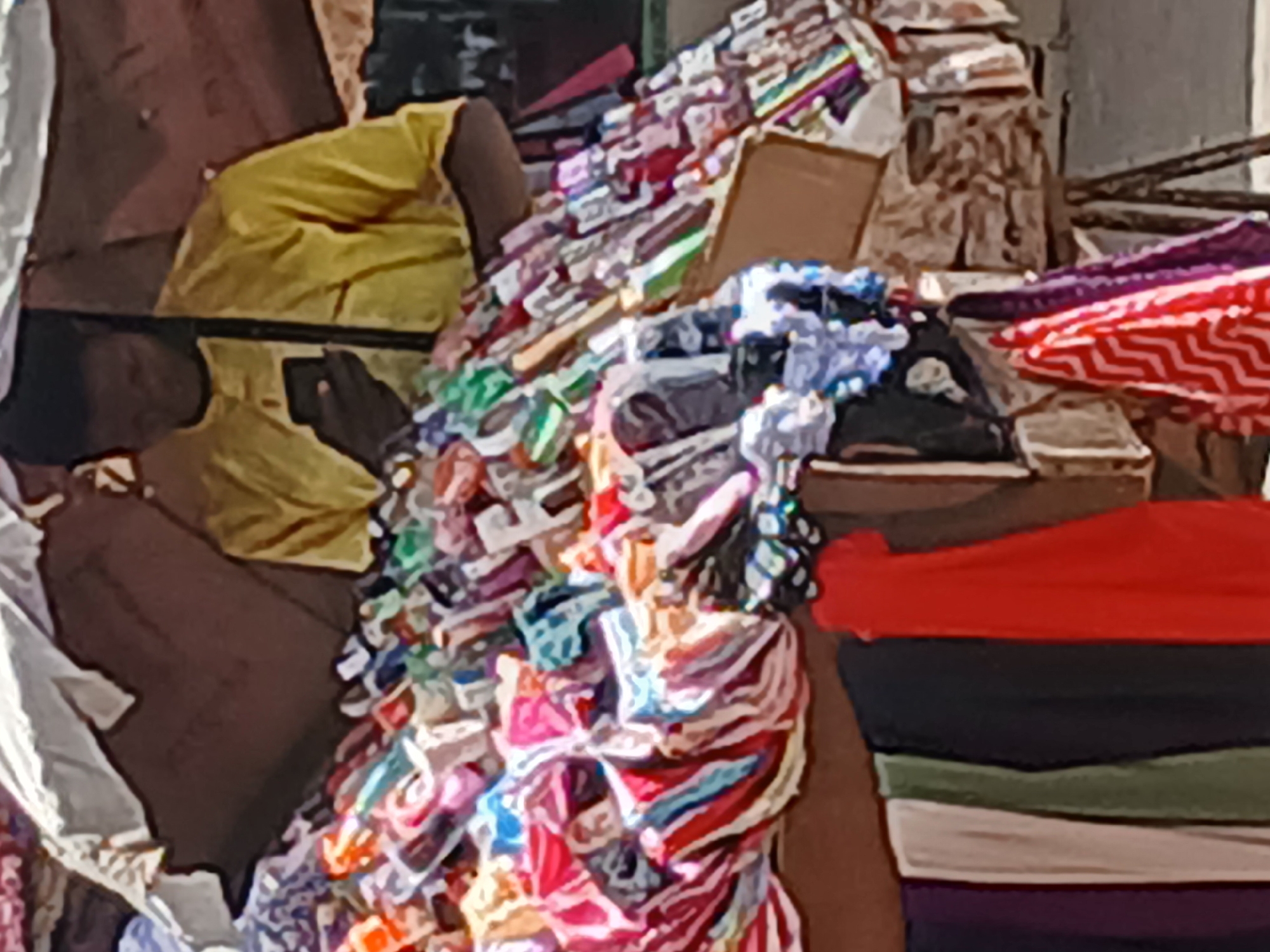 This image depicts a bustling market scene in Maiburiji, Nigeria, focusing on a stall laden with textiles and clothing. A person, partially visible on the left, is wearing a bright yellow short-sleeved top and appears to be seated or kneeling amidst the merchandise. Their dark-skinned hands are visible, with one hand holding a dark rectangular object, possibly a mobile phone.
The scene is filled with a dense array of colorful fabric and clothing items. In the center, a large, jumbled pile of brightly patterned textiles, featuring various hues of red, blue, green, and white, dominates the mid-ground. To the right, a neatly stacked pile of folded garments is visible, showcasing solid colors such as red, black, olive green, white, and purple. Further to the right, items with distinctive red and white zigzag patterns are also part of the display.
The background shows more merchandise, including what might be packaged goods and additional fabric piles, with a cardboard box discernible among them. The abundance and arrangement of the goods suggest an active market or informal retail environment. The lighting indicates it is daytime. No discernible text is visible anywhere in the image. The person's posture and interaction with the items imply they are either organizing or attending to the stall's wares.