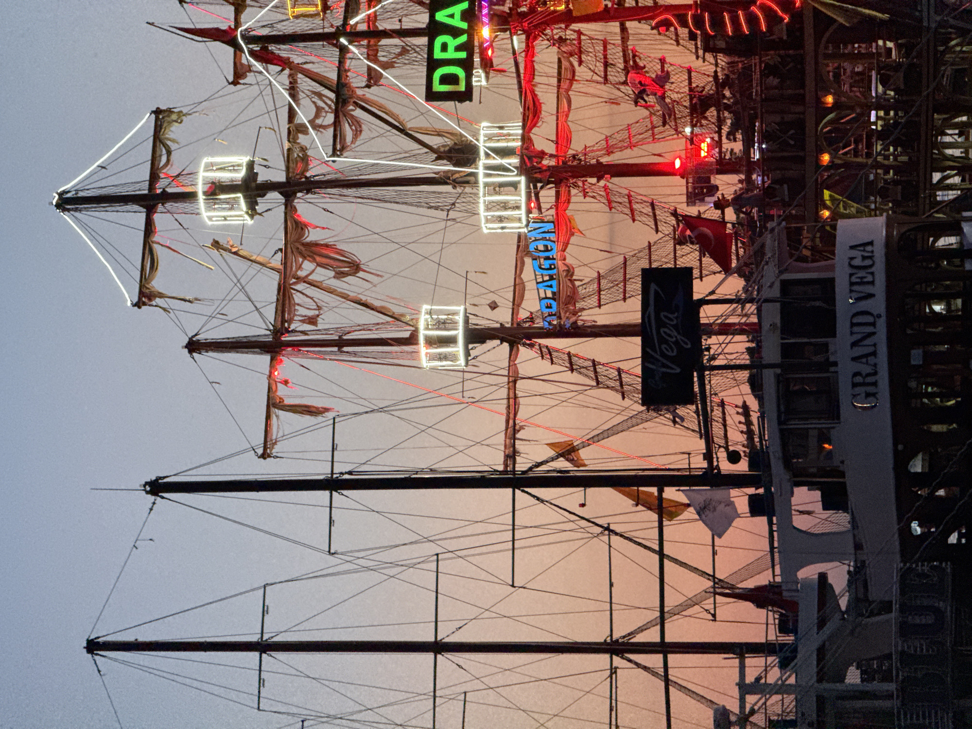 The image depicts a stylized pirate ship, illuminated with neon lights. Visible signage includes the words "DRAGON", "VEGA", and "GRAND VEGA". The ship appears to be a tourist attraction or entertainment venue. The presence of a Turkish flag suggests the location is in Turkey. Given the provided information, it is likely the scene is in Fethiye, Turkey.