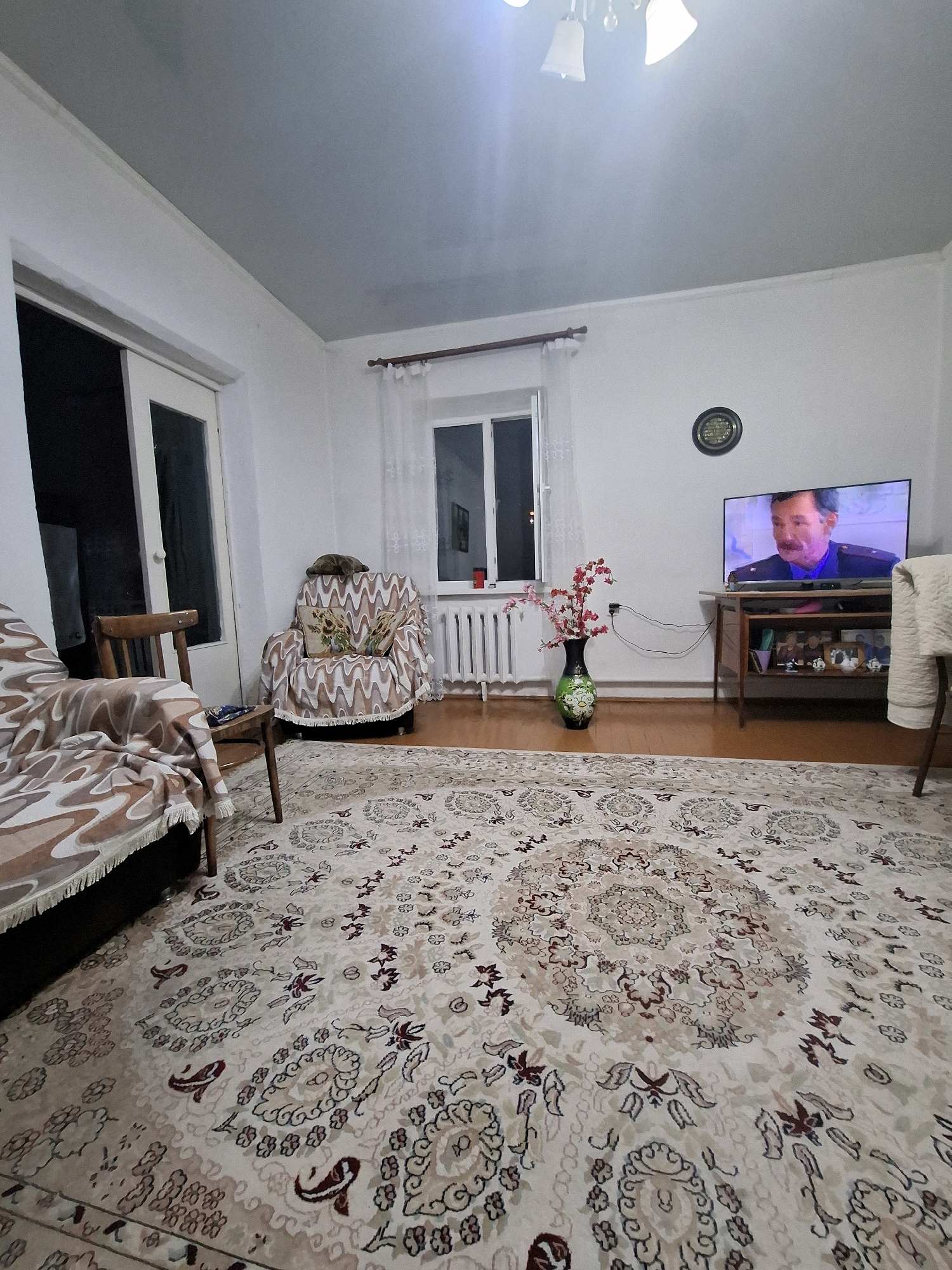 A wide shot captures a living room in Almaty, Kazakhstan. The room is furnished with a sofa, an armchair, and a television on a stand. A large, ornate rug covers the floor. A window with sheer curtains is visible, as is a sliding glass door. The walls are white, and the ceiling is a light gray. A chandelier hangs from the ceiling. A clock on the wall shows the time as approximately 2:40. A vase with artificial pink flowers sits on the floor next to a radiator. The overall impression is of a lived-in, comfortable space.