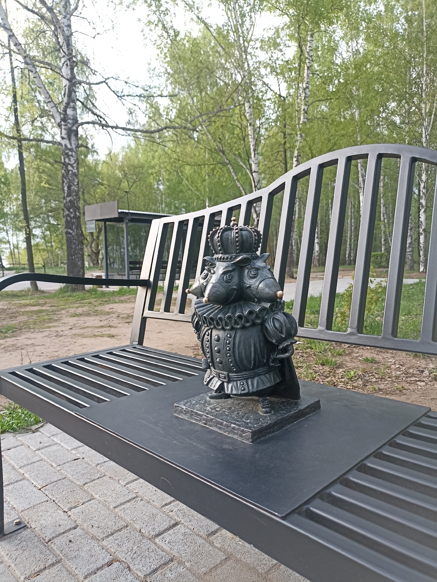 The image shows a metal sculpture of a two-headed rodent wearing a crown and ornate clothing, sitting on a metal park bench. The bench is situated in an outdoor park setting, with birch trees visible in the background and a sheltered bus stop structure visible in the distance. The foreground shows paving stones under the bench.