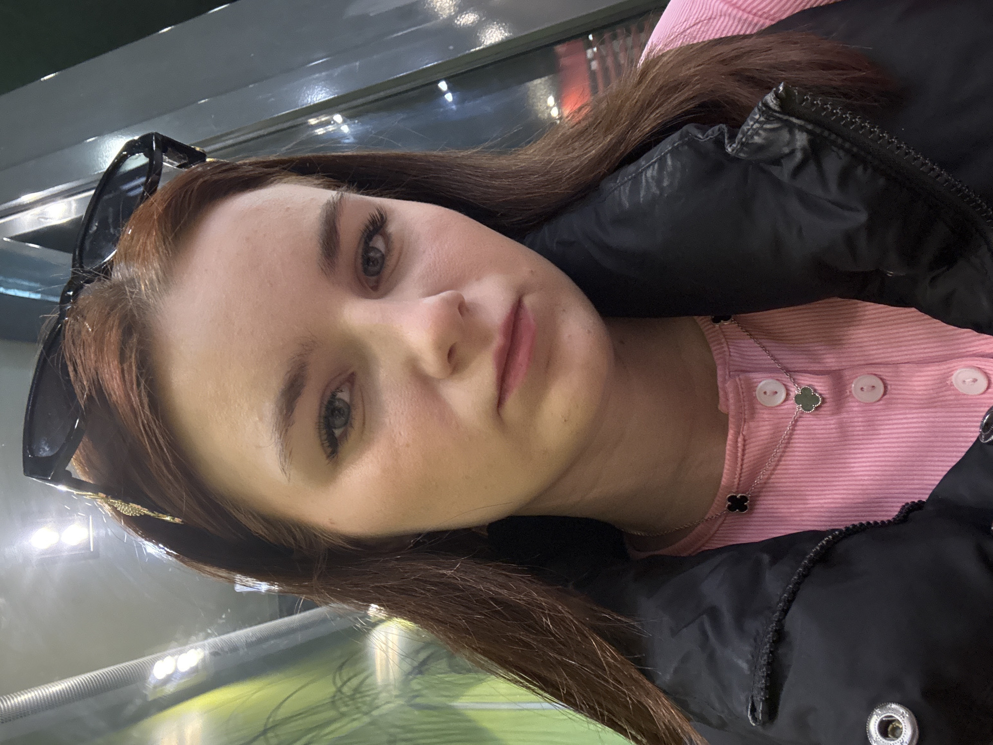 A selfie-style photograph shows a woman with light skin, light eyes, and long brown hair. She is wearing a pink shirt with white buttons, and a black puffy jacket. Sunglasses are perched on top of her head. A silver necklace with a clover-shaped pendant is visible around her neck. The background is blurred, suggesting an indoor environment, possibly a building with glass windows and lighting. There is no information from the image that can confirm this image was taken in Sinyavino, Russia. The photograph primarily focuses on the woman's face and upper body.