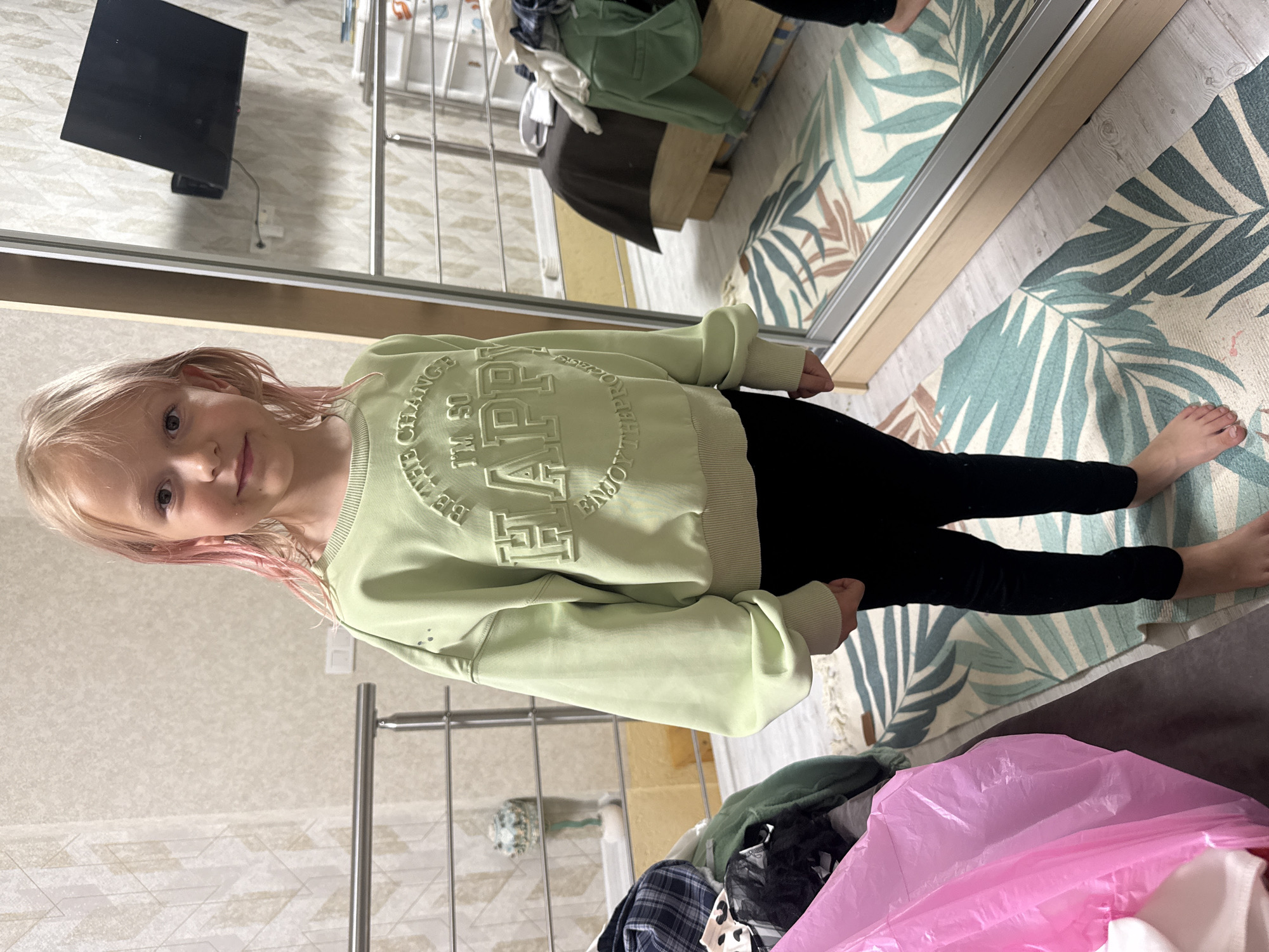 A young girl with blonde hair, tinged pink at the ends, stands in front of a mirror, her body angled away from the camera. She is wearing a light green sweatshirt with raised lettering that reads "BE HAPPY I'M SO". Her right hand is resting on the sweatshirt, and her left arm is bent at the elbow, with her hand near her face. She is wearing black leggings. Her bare feet are visible, with her right foot slightly in front of her left. She is standing on a rug with a tropical leaf pattern in shades of green, teal, and brown. To the left of the rug, the floor appears to be light wood laminate.
In the background, a large mirror reflects a room. A television is mounted on the wall in the upper left corner of the mirror's reflection. A metal railing, likely part of a closet system or room divider, is visible in the reflection, with some clothing items draped over it. A bed with a dark brown comforter is also visible. A portion of a shelf with various items is seen in the reflection.
The scene appears to be indoors, possibly in a bedroom or dressing area. The lighting suggests it is daytime, with no clear indication of the weather. The overall mood is casual and domestic.