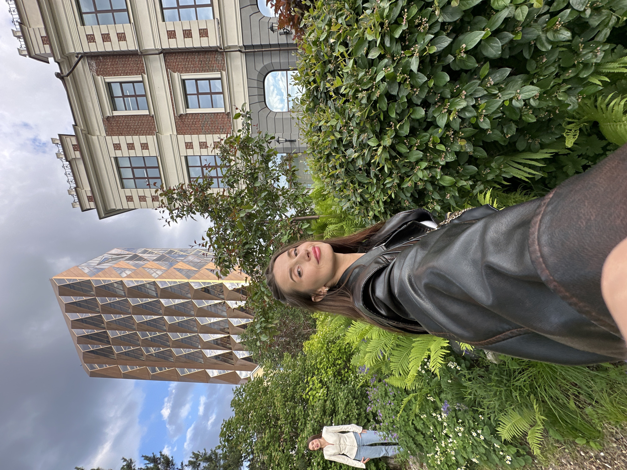 This image, when correctly oriented, captures two young women in an outdoor urban setting against a backdrop of varied architecture and lush greenery.
The primary subject, positioned on the right side, is a young woman with long, straight dark brown hair, wearing a black leather jacket over a dark top. She looks directly at the camera with a gentle smile and her head slightly tilted. Her makeup is subtle, featuring a hint of pink lipstick.
Further in the background, towards the center-left, another young woman is visible. She has lighter-colored hair and is dressed in a light, possibly cream-colored, long-sleeved top and light blue jeans. She stands with her hands in her pockets, looking towards the camera in a relaxed stance.
The immediate surroundings are rich with diverse greenery, including dense bushes adorned with small light-colored flowers or berries, and vibrant green ferns. A small tree with scattered leaves is also present.
In the background, two distinct buildings stand out. To the left, a modern building features a unique geometric facade composed of numerous triangular panels, possibly glass or reflective material, creating a complex, faceted appearance. To the right, a more traditional multi-story building is visible, constructed with red brick and featuring contrasting light-colored architectural trim around its rectangular windows, as well as an arched window on an upper level.
The sky is partly cloudy, displaying patches of blue amidst an otherwise overcast setting, indicating it is daytime. The scene suggests a landscaped area or park within a city, specifically Yekaterinburg, Russia. There is no visible text in the image.