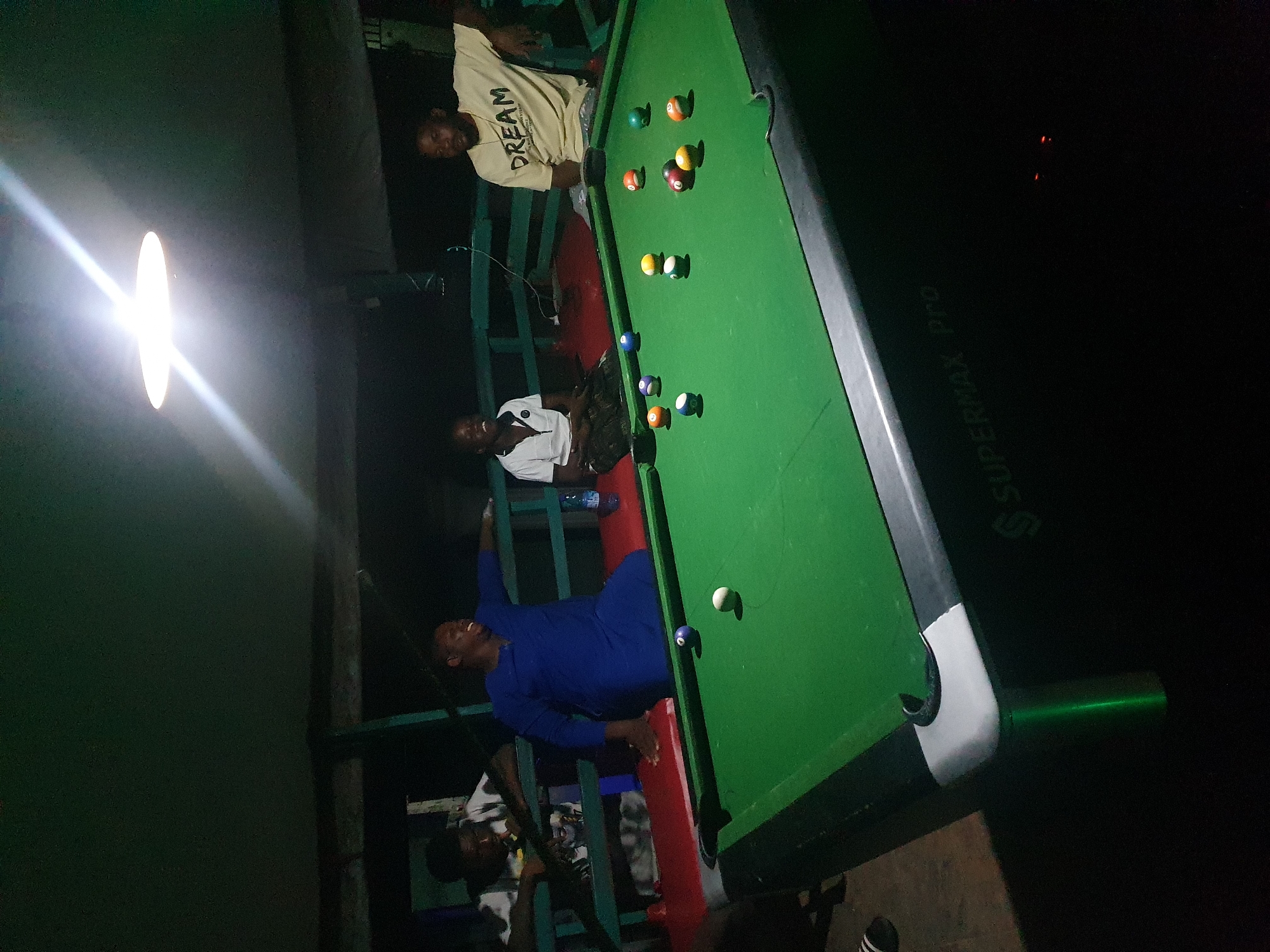 The image depicts an indoor pool hall in Lokoja, Nigeria, where four individuals are gathered around a pool table. The lighting is dim, with a bright overhead light illuminating the scene. Two men are seated on a bench, while another man and a boy stand near the table. The pool table itself is green with various colored balls scattered across its surface. The background shows a dark, enclosed space, possibly a bar or recreational area. The overall atmosphere suggests a casual gathering.