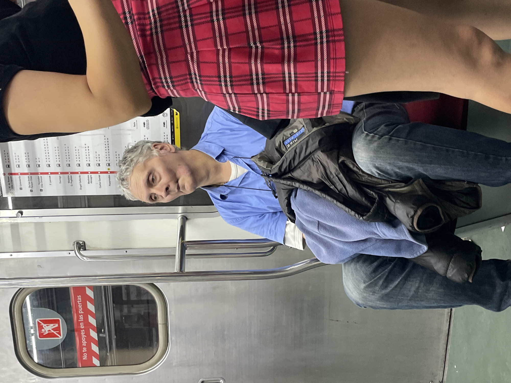 A photograph shows a man sitting on a subway train in Buenos Aires, Argentina. The subway car has a sign that says "No te apoyes en las puertas." A subway line map is visible above the man's head. The man is seated, wearing a blue shirt and jeans. A jacket is draped over his lap. An arm and a red plaid skirt are visible in the top portion of the image. The window has a sign that says "No te apoyes en las puertas," and the name "Villa Urquiza" is visible on the subway line map. The name "Almagro" is also visible on the subway line map.