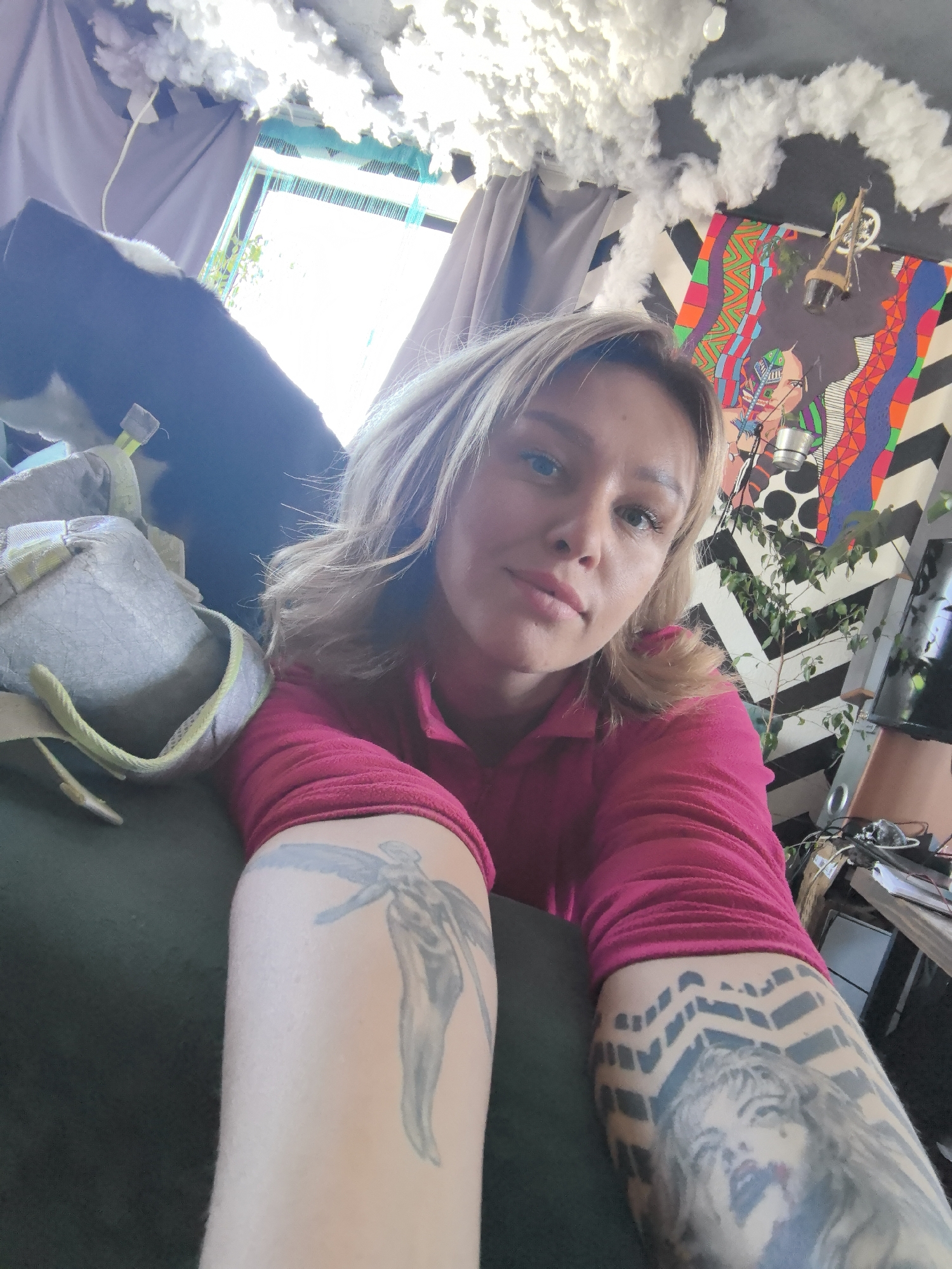 An adult female is depicted in a close-up selfie, positioned indoors. She has blonde hair and fair skin, looking directly at the camera with a neutral expression. She wears a long-sleeved fuchsia-colored top. Her right forearm (viewer's left) displays a grayscale tattoo of an angelic figure with wings. Her left forearm (viewer's right) features a tattoo of a woman's portrait with red lips, surrounded by a black chevron pattern. She appears to be leaning against a dark green or black upholstered surface, possibly a couch or bed, where a gray bag with light green accents is also visible to her left.
The background reveals an eclectic indoor environment. A wall behind her features a large, colorful, abstract geometric mural composed of various patterns and shapes. Below this mural, the wall is covered with a black and white chevron patterned material. Small plants in hanging containers are affixed to the wall near the mural. To the upper right, the black ceiling is decorated with numerous white, cloud-like structures. A brightly lit window is visible in the upper left, partially obscured by dark curtains, with a hint of green foliage outside. To the lower right, a metallic object resembling a ladder and some visible wires suggest a work or storage area. The specific city of Noviy, Russia, cannot be determined from the visual information in the image.