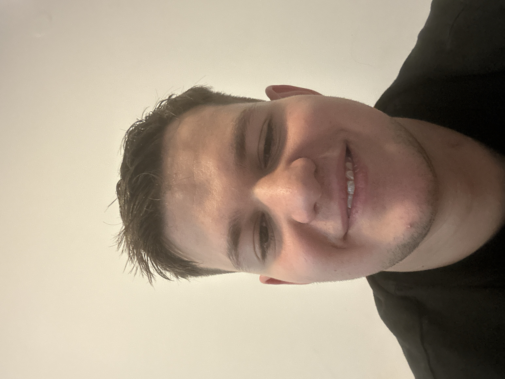 A close-up portrait of a smiling young man with short, dark hair. He is wearing a black shirt. The background is a plain, light-colored wall. There is no indication of location within Sofia, Bulgaria, or any other city. The focus is solely on the individual.