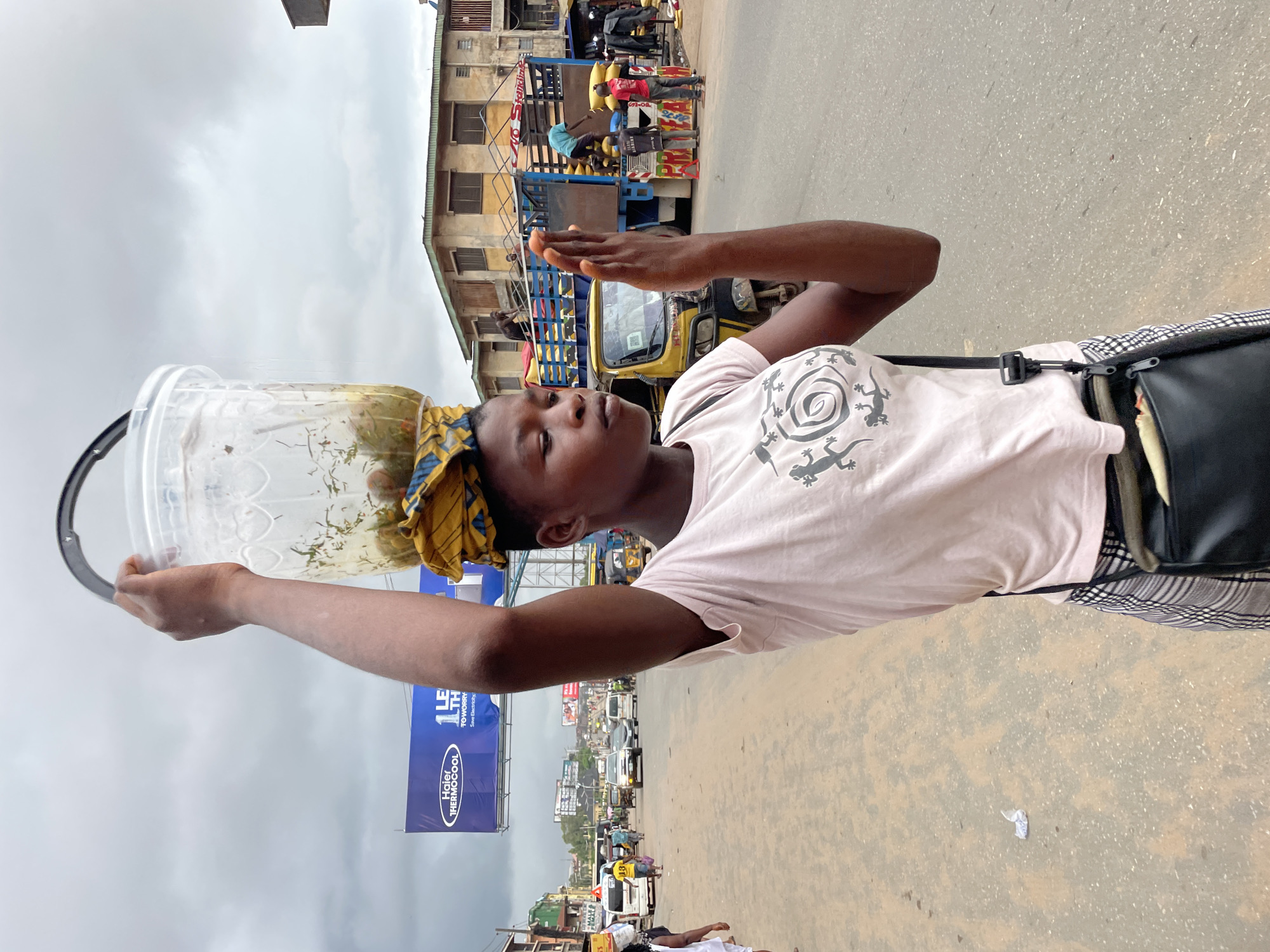 The image depicts a young woman carrying a transparent plastic bucket filled with food items on her head, walking on a dusty, paved street in Onitsha, Nigeria. She wears a pale pink t-shirt with a gecko-like graphic and a checkered skirt. A black crossbody bag is slung over her shoulder. Her right hand is raised, palm outward, as if signaling or interacting with someone. In the background, traffic including cars, a yellow tricycle, and a truck laden with goods suggests a busy urban environment. Buildings line the street, and a large blue billboard for "Haier Thermocool" is visible. The sky is overcast. The scene appears to be a typical market or street scene in a Nigerian city, likely depicting a vendor or carrier of goods.