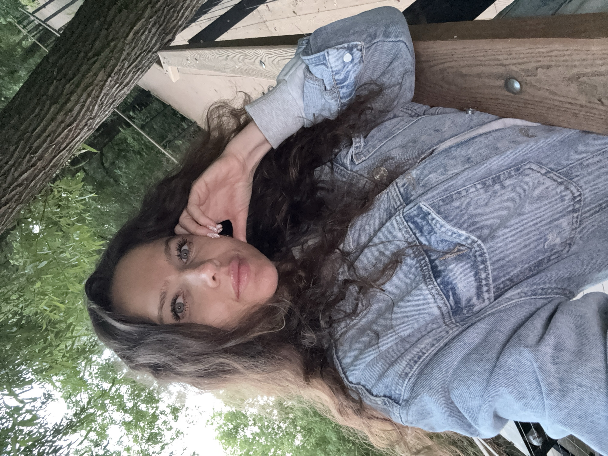 A woman with long, wavy brown hair and blue eyes is captured in a selfie. She is wearing a light blue denim jacket over a grey shirt. Her right hand is resting on her cheek, with her fingers touching her face. The background shows lush green foliage and a wooden railing, suggesting an outdoor setting. The lighting indicates it is daytime, possibly late afternoon or early evening. The image is taken from a medium close-up perspective, focusing on the woman's face and upper body. The specific location in Moscow, Russia, cannot be determined from the provided image alone.