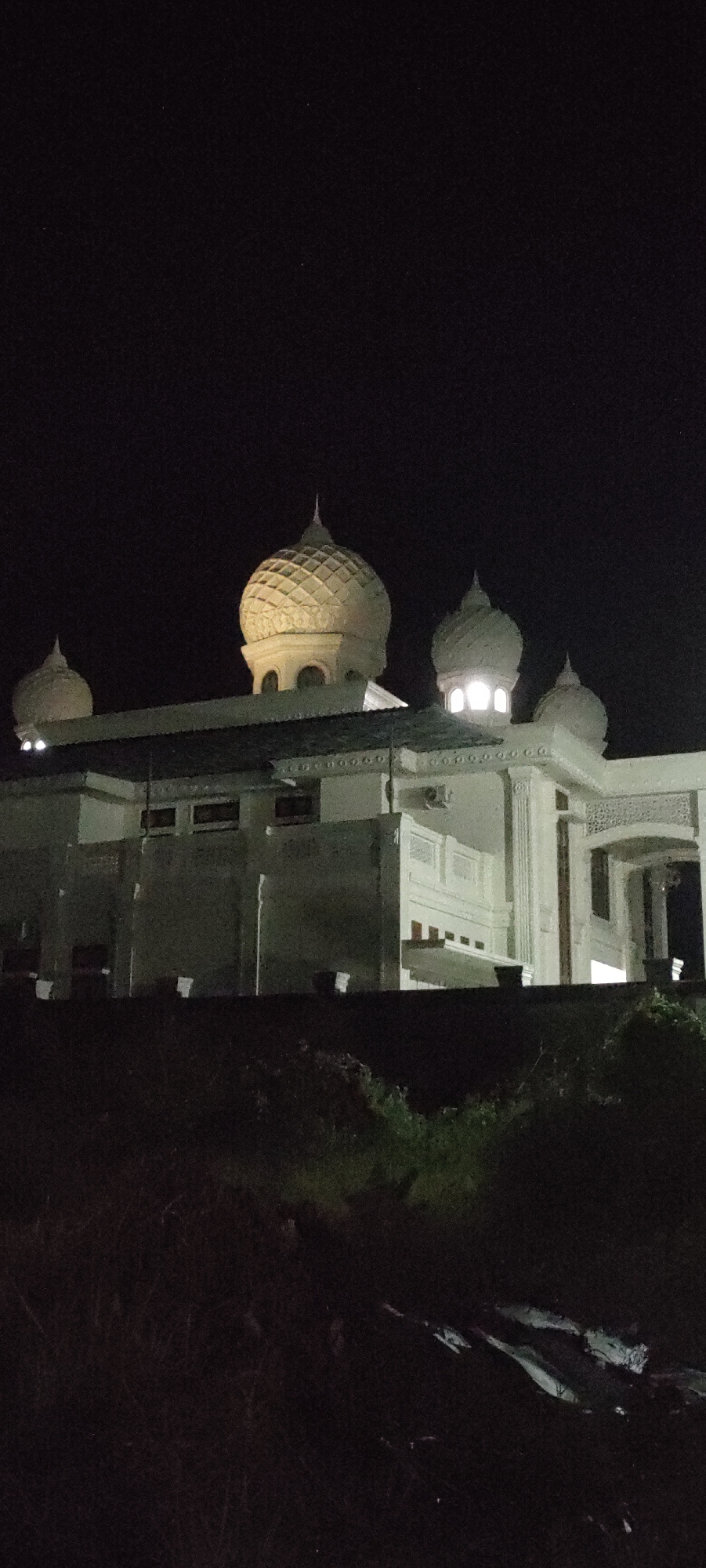 At night, a view of the Islamic Center Mosque in Lhokseumawe, Indonesia. The building is white and has several domes on top, one of which is large and patterned. Illumination emanates from the dome windows. There is some vegetation in the foreground.