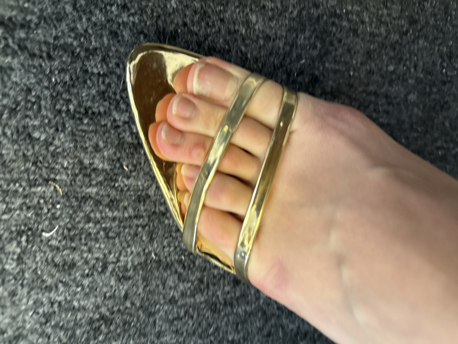 A single, fair-skinned human left foot is depicted wearing a gold-colored, open-toed sandal. The scene is located in Arcueil, France. The sandal features a metallic finish with two parallel, reflective straps securing the forefoot and toes. The toes display natural toenails with unpainted, visible white tips. Minor dermal irregularities, consistent with calluses, are present on the superior surface of the second and third toes. The foot and sandal rest on a dark grey, densely textured carpet. Sparse, light-colored fibrous debris is visible on the carpet in the background.