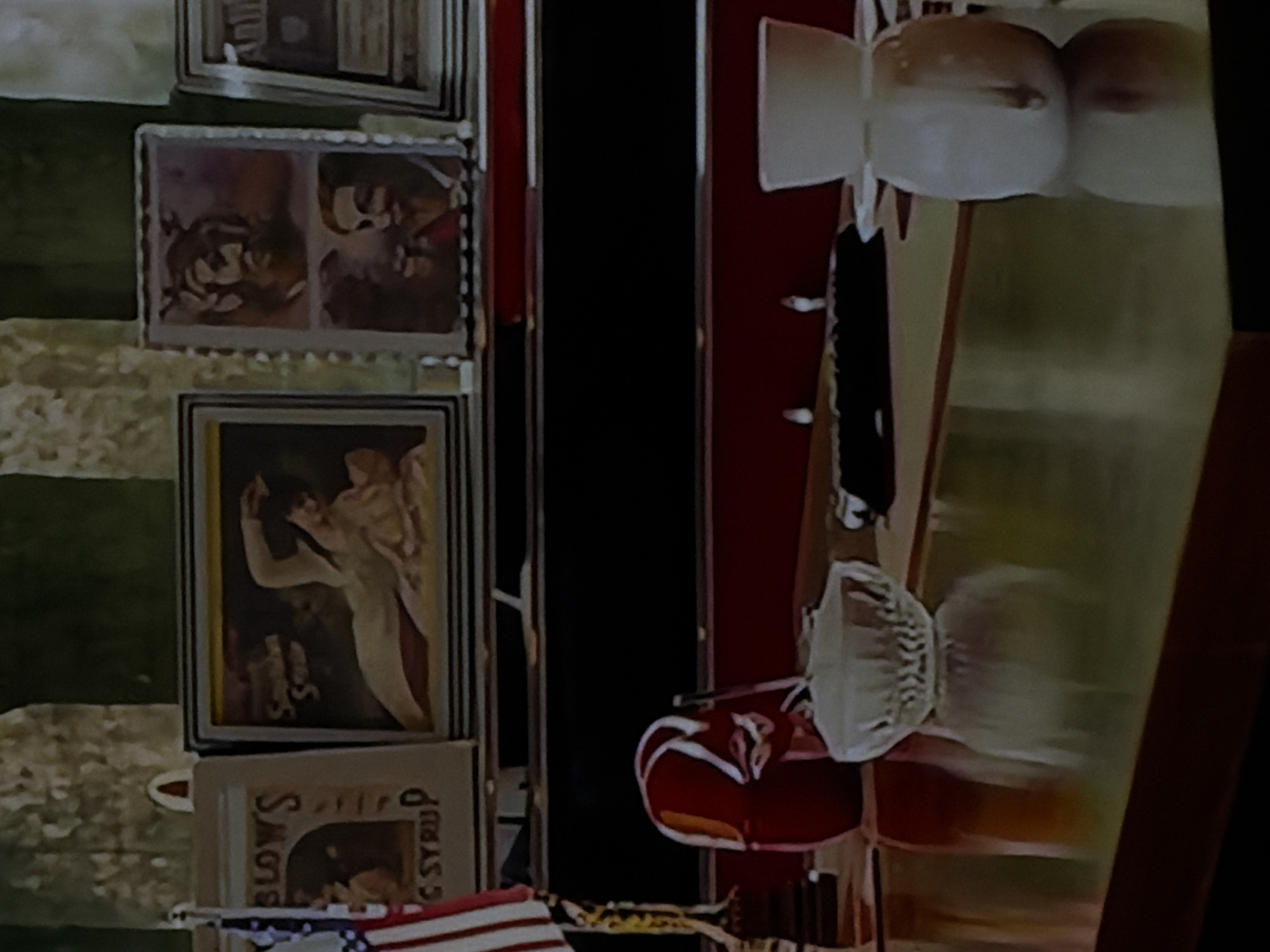 The image captures an indoor scene, possibly a room or a commercial space, decorated with various items. The perspective is slightly angled, looking across a room.
On the left side, a green wall or patterned wallpaper serves as a background for several framed pictures or posters. From top to bottom:
* The topmost visible frame is partially obscured.
* Below it, a rectangular frame contains two distinct images side-by-side. The left image shows a figure with a prominent beard or facial hair, possibly a caricature or stylized portrait. The right image depicts another face, also with prominent facial features.
* Further down, another framed image features a light-skinned figure, possibly a woman in an elegant, flowing garment, looking upwards. Text "Cream Soda" is partially visible in the bottom left corner of this artwork.
* The lowest visible frame contains an old advertisement. The words "BLOWS" are clearly visible at the top, and "C SYRUP" (likely "CORN SYRUP") can be seen at the bottom right. A small American flag, striped red and white, is visible beneath this frame, suggesting a patriotic or American-themed decor.
To the right of these framed items, a dark, vertical gap separates the left wall from the right side of the image, which features furniture or display elements. The right side is dominated by red surfaces, possibly part of a cabinet, counter, or booth seating. On a horizontal surface, possibly a shelf or table, several objects are placed. These include what appear to be light-colored mugs or small bowls, arranged near the top right. Further down, a decorative red helmet with white accents, resembling a baseball helmet, is visible. Another patterned, light-colored object, possibly a bowl or dish, is also on this surface.
The overall impression is one of vintage or retro decor, with a potential American influence given the flag and old advertisements, set in Sofiivska Borshchahivka, Ukraine. The lighting is artificial and consistent with an indoor setting.