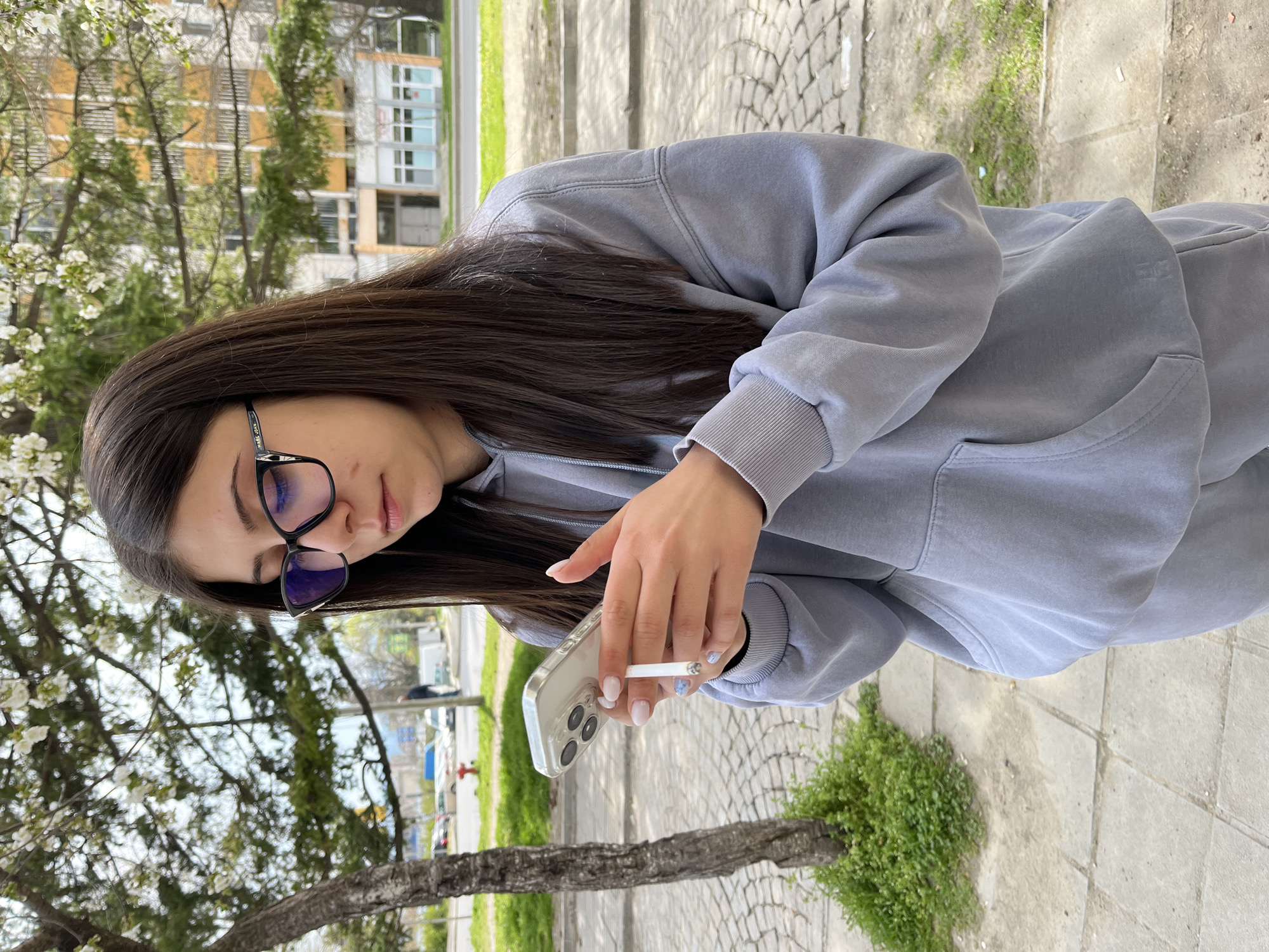 A young woman stands outdoors, angled towards the viewer, appearing to be in Karlovo, Bulgaria. She has long, dark brown hair and wears glasses. The woman is dressed in a light gray hoodie and matching sweatpants. She is holding a smartphone in her left hand and a lit cigarette in her right hand. The background includes trees with white blossoms, a building, and a cobblestone walkway. The location is outdoors during the daytime.