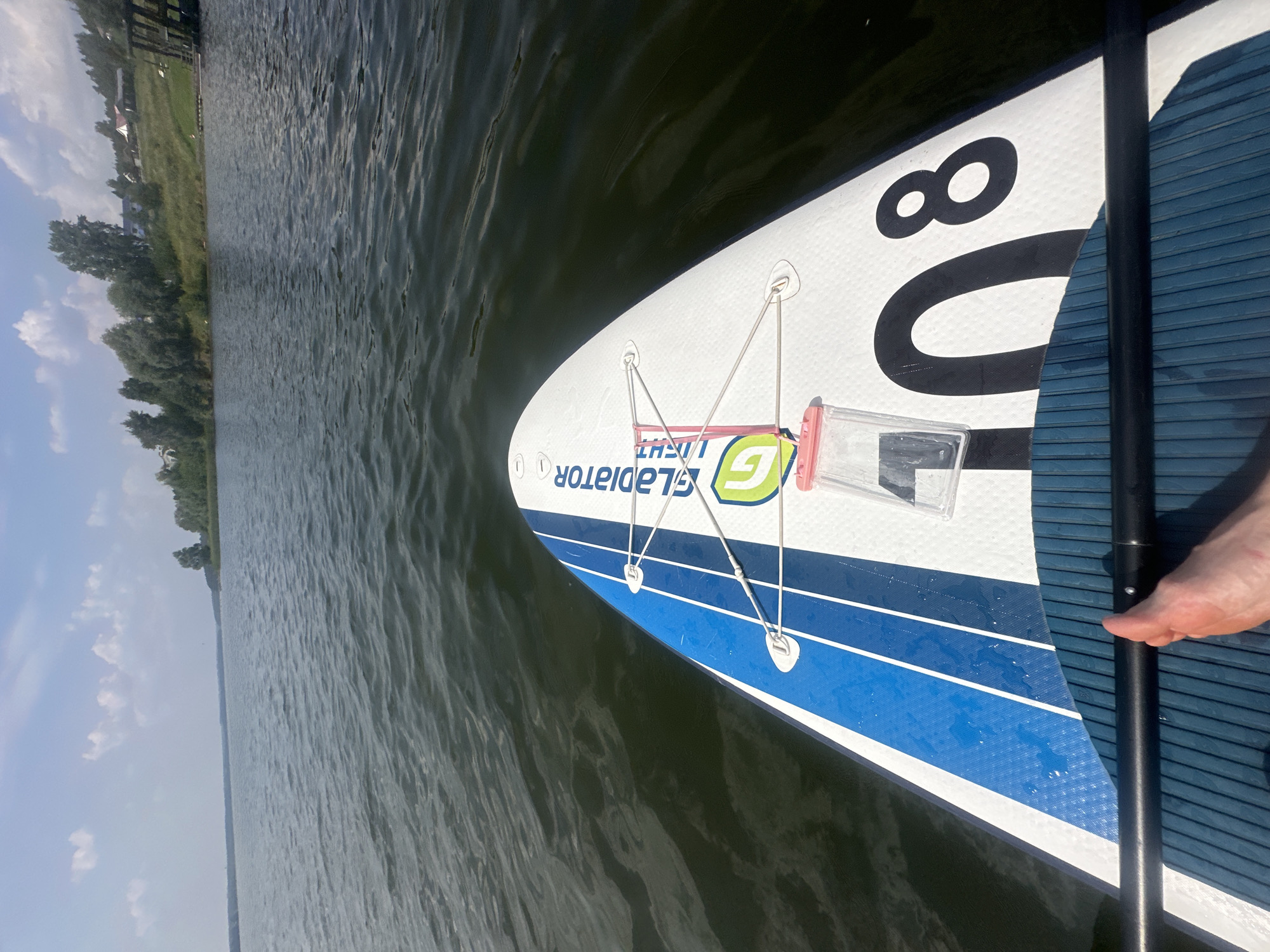 The image shows a first-person perspective of someone on a paddleboard in the water. The paddleboard is white with blue stripes and the number "08" in black is visible on the tail. A black paddle is resting on the board, and a foot is visible near the paddle. A clear waterproof pouch with a pink closure is secured to the board with elastic cords. The background shows a calm lake with a tree-lined shore and some buildings on the distant bank. The sky is partly cloudy. The scene appears to be in Votkinsk, Russia, given the context of the provided location.