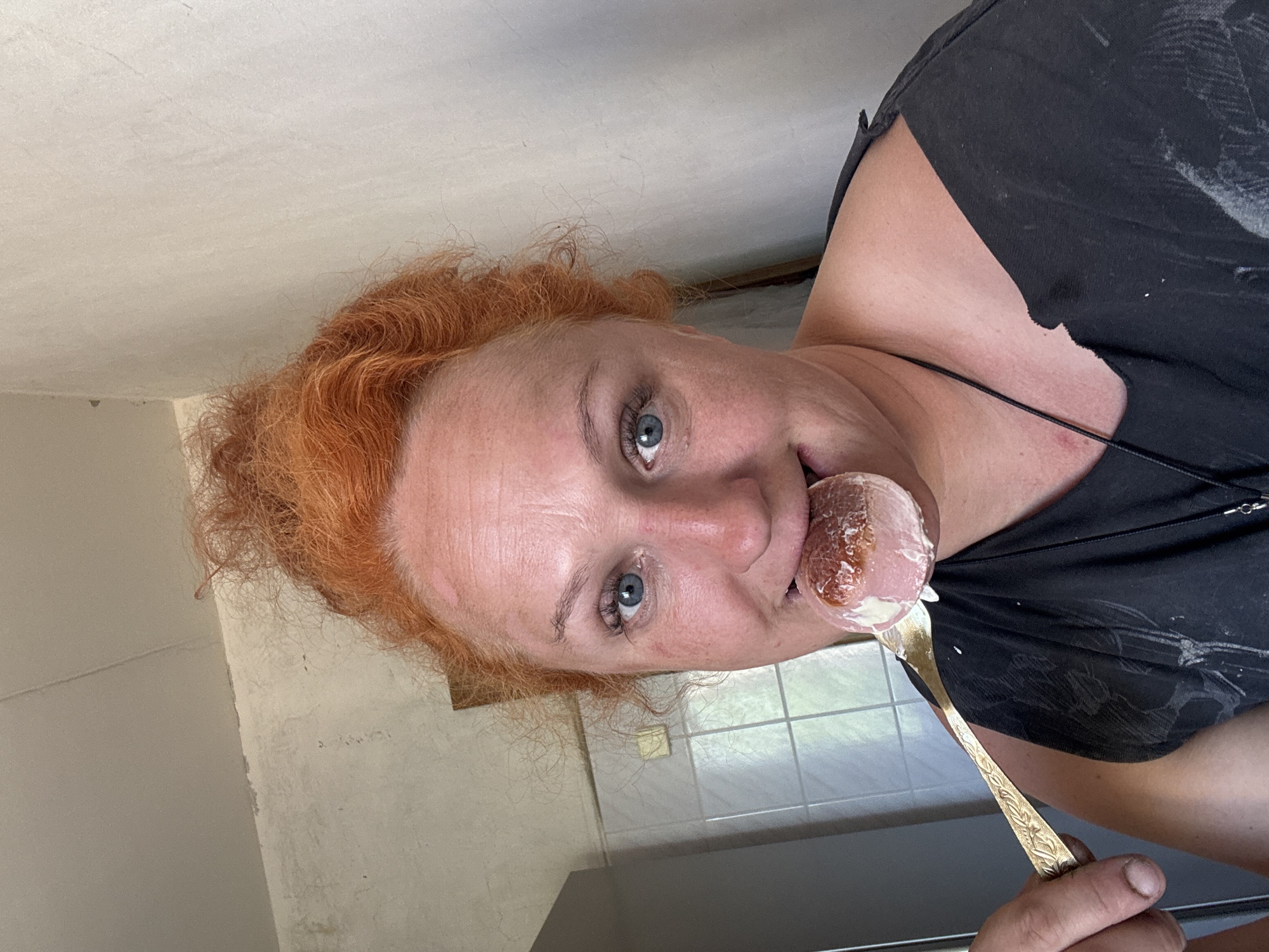 A fair-skinned person with red hair is holding a spoon with food in their mouth. The person is wearing a black shirt. The spoon appears to be gold in color. The background shows a wall and a tiled area. There is no indication of the city of Chaikovsky, Russia, in the image.