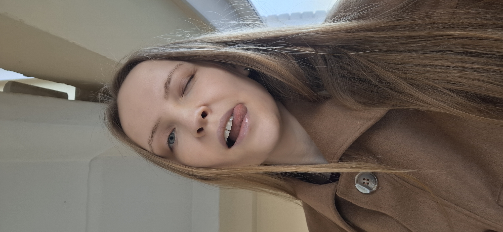 A young woman with long, straight, light brown hair is depicted in a playful, close-up portrait, taken from a low angle. She is winking with her left eye (viewer's left) and has her mouth slightly open with her tongue extended in a mischievous gesture, revealing her upper teeth. Her visible right eye is a light blue, and her skin appears fair and clear.
She is wearing a warm, camel-brown coat, possibly made of wool, with a single speckled, round brown button visible on the right side. The background is simple and bright, consisting of light-colored walls and what appears to be a window or bright light source at the top, suggesting a daytime indoor setting. The overall emotion conveyed is cheerful, lighthearted, and relaxed. There is no visible text. While the location is provided as Chaikovsky, Russia, the image itself does not contain specific visual cues to indicate this geographical setting.