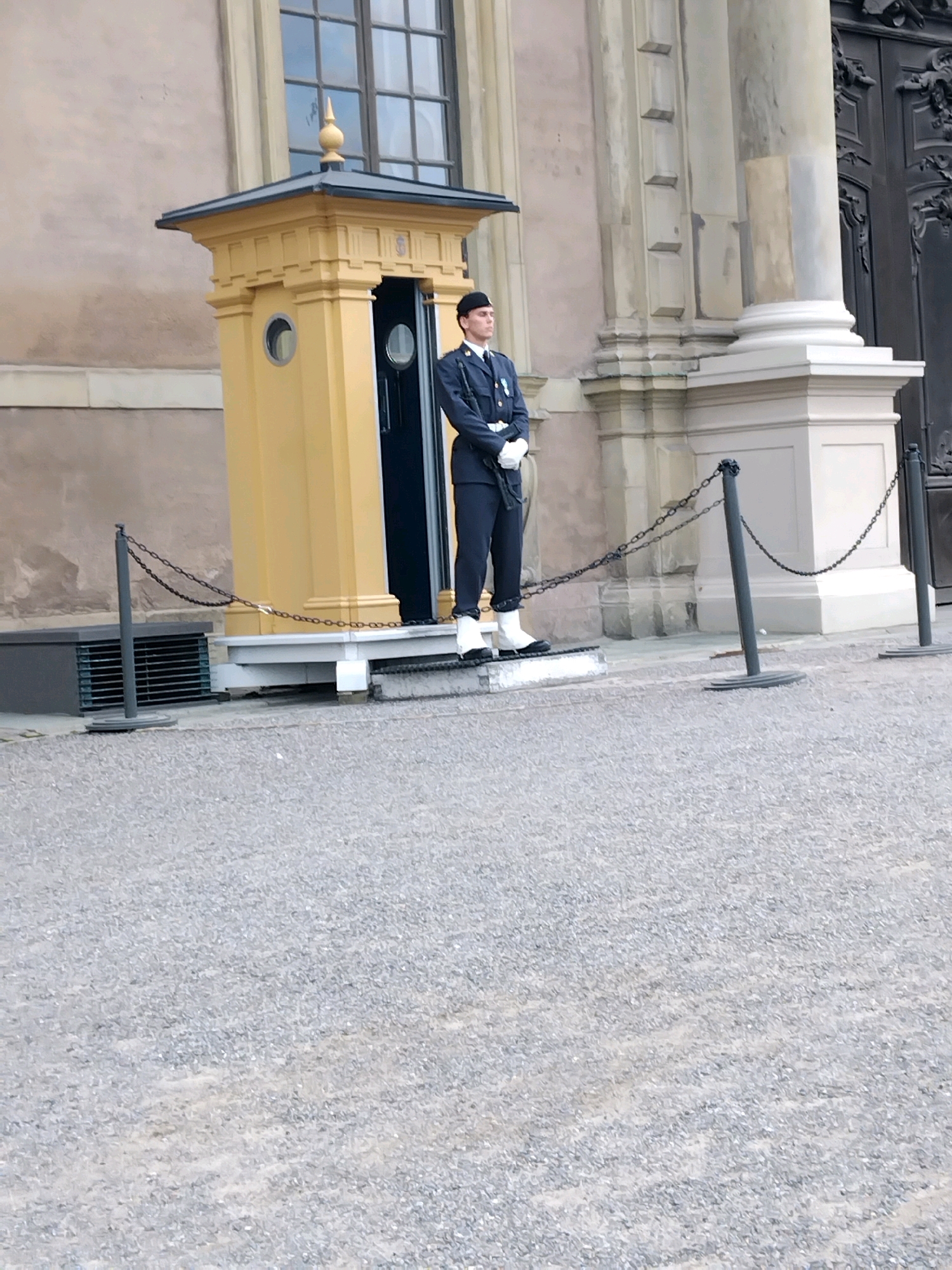 A uniformed guard stands at attention outside a yellow, ornate guardhouse. The guard wears a dark uniform with white gloves, white gaiters, and a black hat. He stands with his hands clasped in front of him. The guardhouse has a dark doorway and a decorative finial on its roof. A chain barrier is in front of the guardhouse. The scene takes place in front of a large building with classical architectural elements, including columns and a large window with multiple panes. The ground is covered in gravel. This scene is located at the Stockholm Palace, specifically in the Outer Courtyard.
