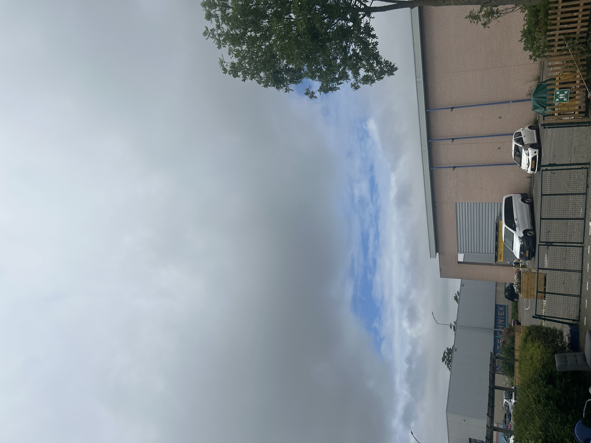 The image shows an outdoor scene with a building and a cloudy sky. The building appears to be a commercial or industrial property with a brick facade and large windows or doors. There are several vehicles parked outside, including a white car and a white van. A wooden fence and some greenery are also visible. A sign with green arrows indicating directions for people is posted on the fence.
The sky is mostly cloudy, with some patches of blue sky visible. The lighting suggests it is daytime, possibly late afternoon or early evening. The scene seems to be located in an urban or suburban area.
Visible text includes "DHL" on the side of the white van. There is also some blue text on the side of a building that appears to read "APPSTECHNIEK".