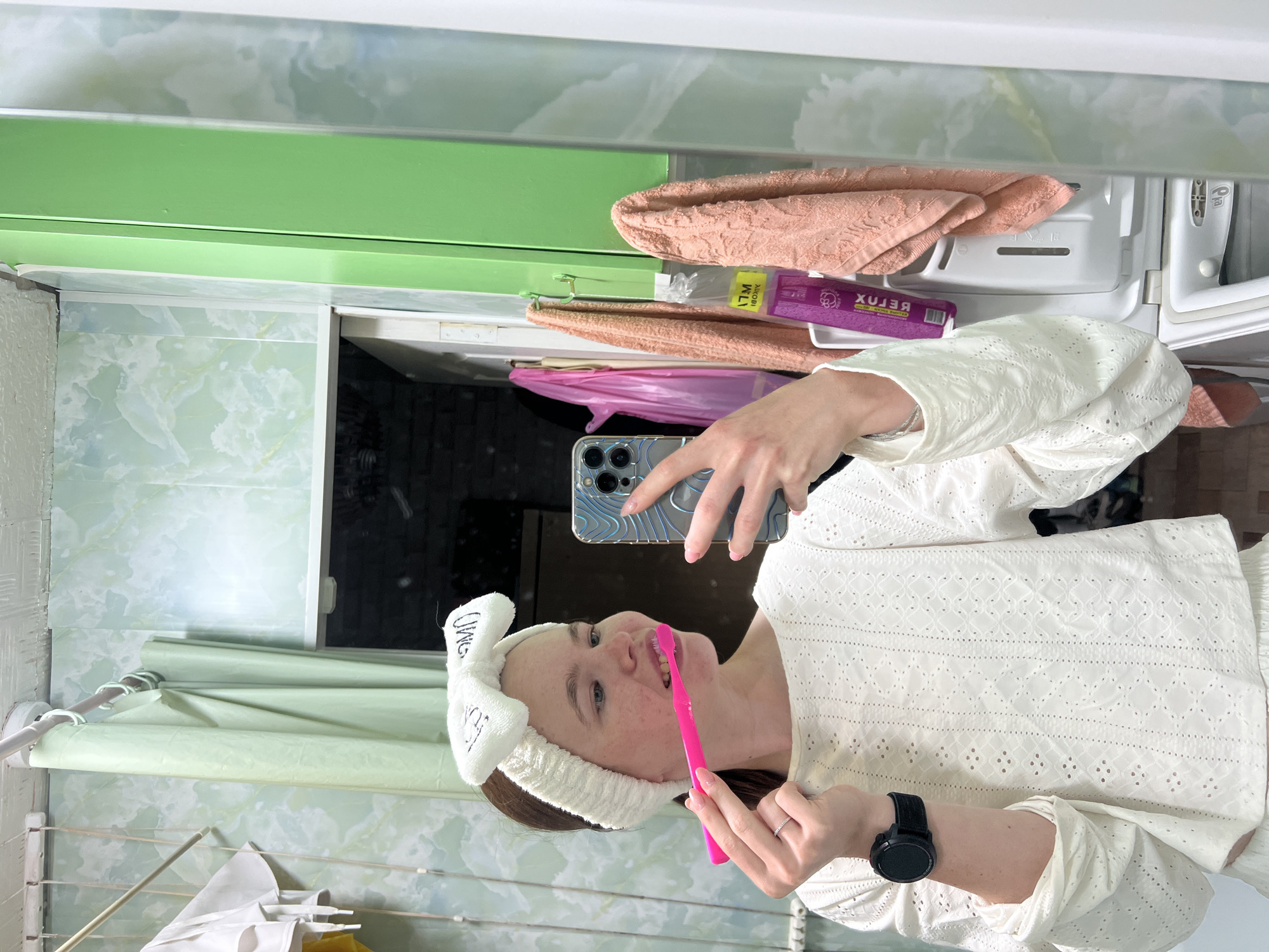 A woman is standing in a bathroom, facing a mirror, and taking a selfie while brushing her teeth. She is wearing a white headband with bunny ears and a white eyelet shirt. She is holding a pink toothbrush in her right hand and a smartphone in her left. A black smartwatch is visible on her left wrist. The bathroom has light green marble-patterned walls and a green shelf above the sink. There are peach-colored towels, and a purple box of "Xuxu" product. Laundry hangs in the mirror's reflection. I can not confirm that the photo was taken in Chaikovsky, Russia.