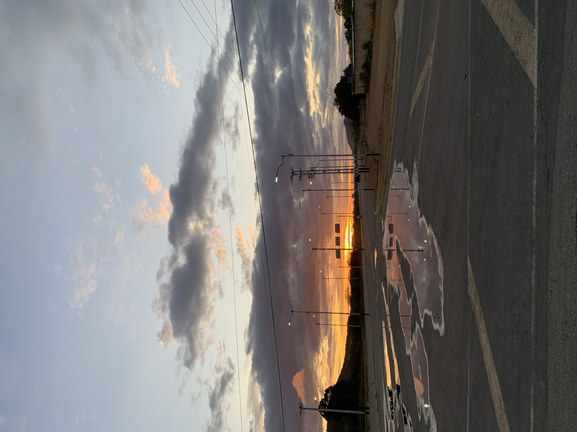 This outdoor landscape photograph, taken in Santa Ana, Venezuela, captures a dramatic sunset or dusk scene. The sky dominates the upper portion of the frame, filled with a mix of dark grey, stormy-looking clouds and luminous patches of orange and yellow, brilliantly lit by the setting sun on the horizon. Lighter grey-blue sky is visible in areas where the clouds are thinner.
Numerous utility poles with interconnected power lines stretch across the mid-ground, silhouetted against the vibrant sky. Several streetlights are also present along the side of what appears to be an asphalt road or parking lot, with some appearing to emit light or reflect the ambient glow. The ground, which occupies the lower half of the image, features dark asphalt with distinct white and yellow painted lines.
A prominent feature of the foreground is a series of puddles on the asphalt, reflecting the rich, warm colors of the sky and the glowing streetlights, creating a mirrored, somewhat abstract view of the scene above. To the right of the road, there is a strip of dry, light-brown earth with sparse vegetation and a low, light-colored wall or barrier. In the distance, a dark, low mountain or hill range is visible beneath the horizon, further defining the setting.
There are no people or discernible activities taking place, contributing to a tranquil and still atmosphere. The image primarily focuses on the interplay of natural light, clouds, and reflections in a semi-urban or rural transition zone, likely after recent rainfall given the puddles. No visible text is present in the image.