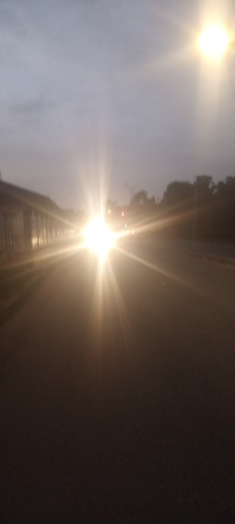 The image shows a road in Kabala, Nigeria, at dusk or dawn. There is a bright light source, likely headlights, in the distance. To the left, a building or structure with arches is partially visible. Streetlights can be seen in the far distance, along with roadside vegetation. The road itself is asphalt and takes up most of the frame.
