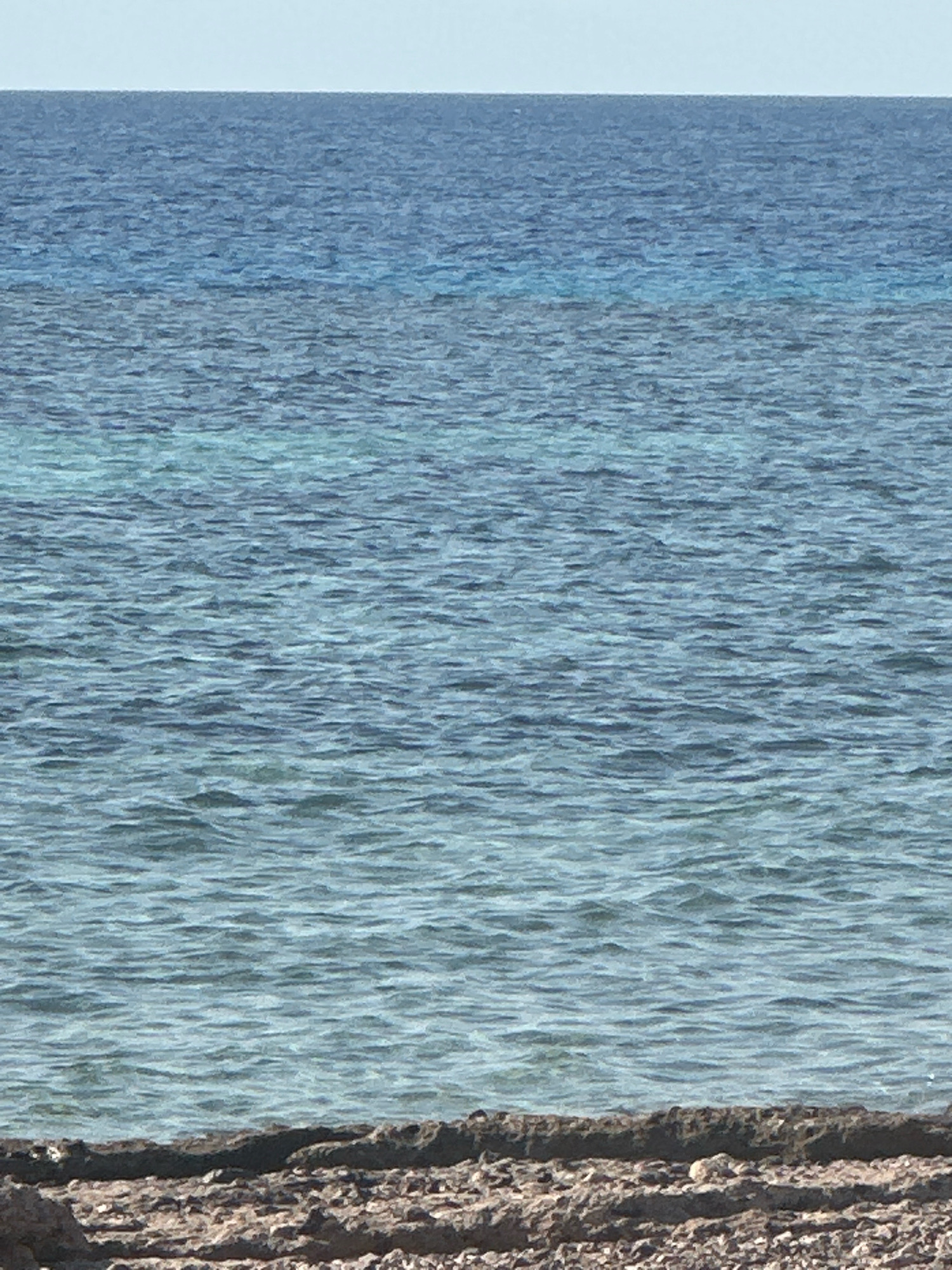 The image shows a close-up view of the sea and a rocky shoreline. The water is a deep blue, transitioning to lighter turquoise hues closer to the shore, indicating a shallow area or possibly a reef. The surface of the water is rippled, suggesting a gentle breeze. The shoreline is composed of rough, dark rocks and pebbles. The sky is a pale, clear blue, almost white at the horizon. The image is taken from a low angle, focusing on the water and the immediate foreground of the shore. The scene is devoid of any people or prominent man-made objects, offering a natural and tranquil coastal landscape. This type of scenery is typical of the Red Sea coast in Nueiba, Egypt, known for its clear waters and rocky shores.