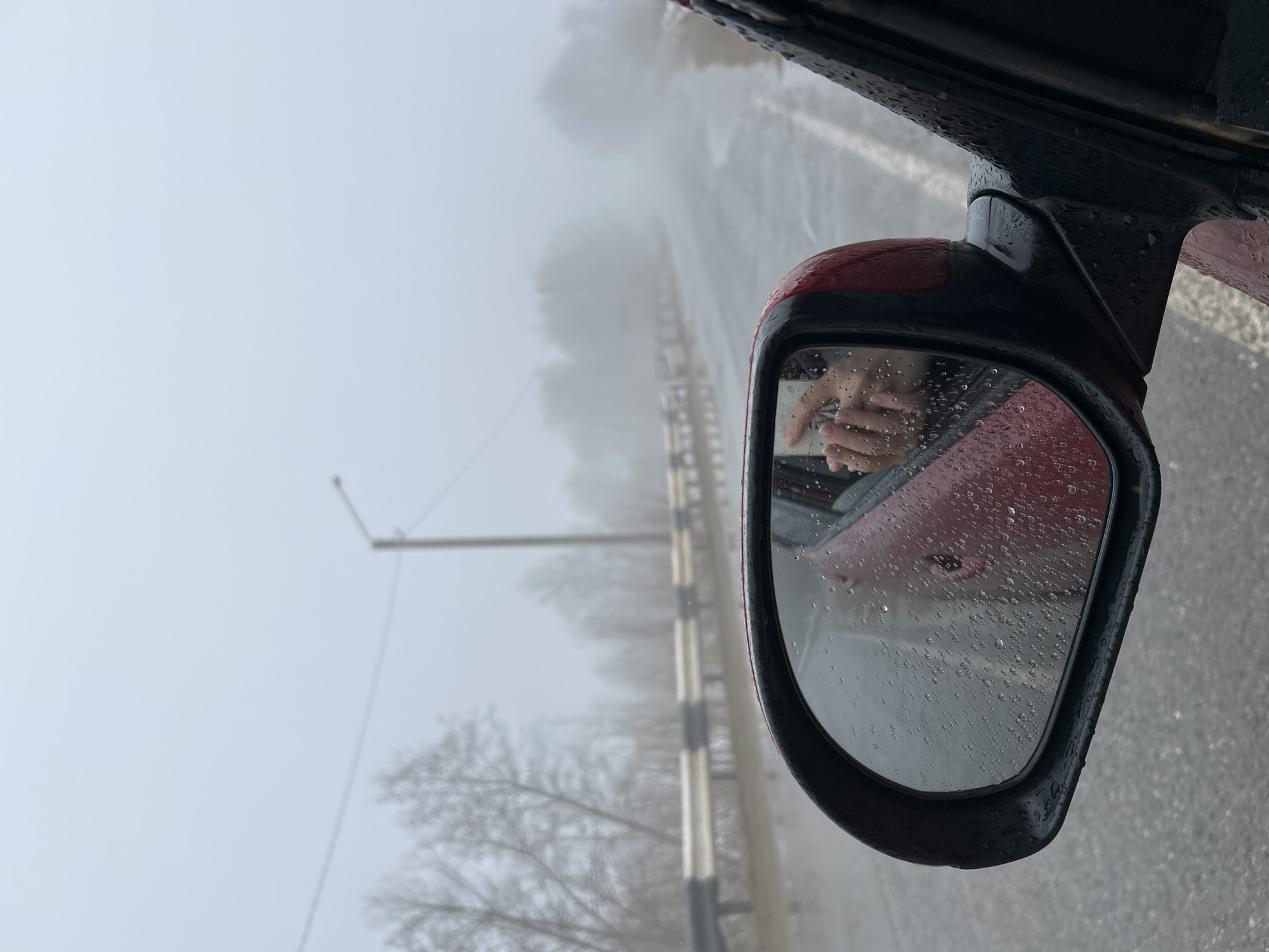 The image depicts a view from inside a red car driving on a road in Уральское, Russia. The weather appears to be overcast and possibly rainy, with low visibility as indicated by the fog. The side mirror of the car is visible, covered in raindrops, and reflecting the interior of the car. Inside the car, a pair of hands is visible in the side mirror. Outside the car, the road is wet and a streetlight and roadside barriers are partially visible through the fog. Bare trees line the roadside.