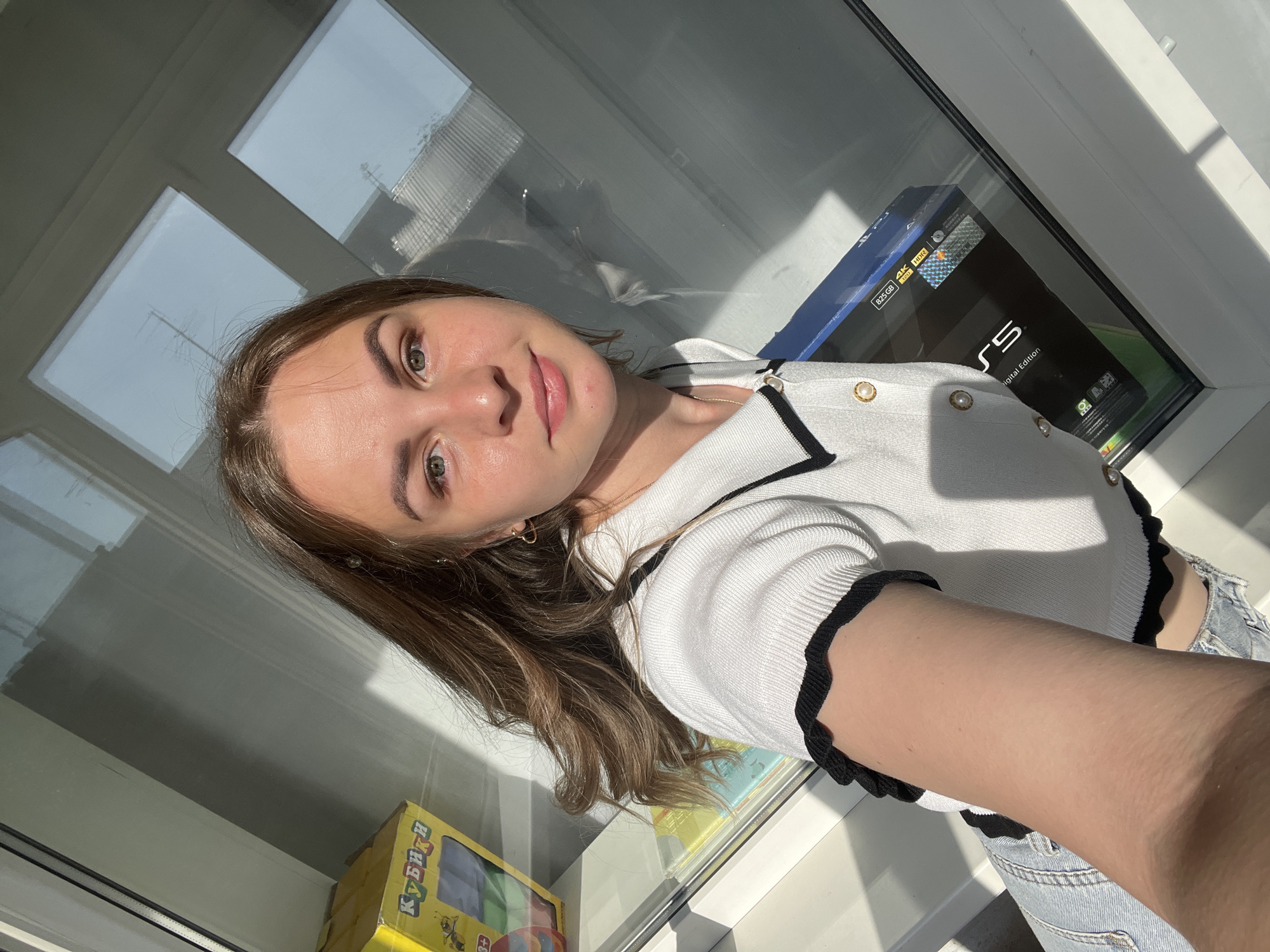 A young woman is taking a selfie indoors, likely in a well-lit room with large windows. She has light brown hair and is wearing a white top with black trim and what appears to be blue jeans. Behind her is a window sill where a PlayStation 5 box and a yellow box with "КУБИКИ" (Cubes) written on it are placed. The scene is presumably in a residential building, with a view of other buildings visible through the window. There is no evidence to definitively confirm the location in the city of Izhevsk, Russia.