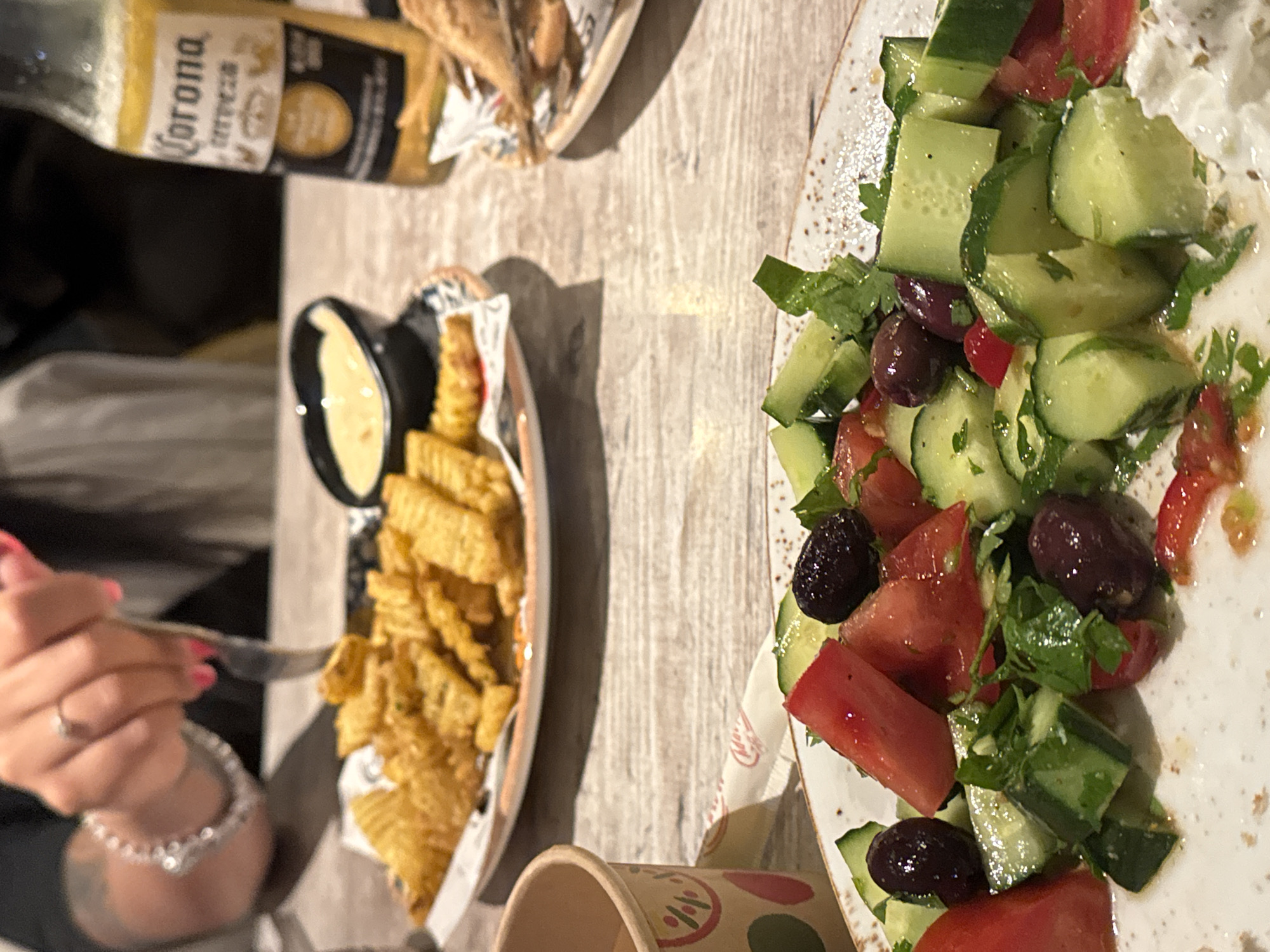 This image captures an indoor dining scene, likely in a restaurant or bar in Sofia, Bulgaria. The setting features a light-colored, wood-grain table laden with food and drink.
In the foreground on the left, a person's hand is visible, holding a fork. The hand has pink nail polish, a ring on a finger, and a sparkly beaded bracelet on the wrist, with a tattoo partially visible on the inner forearm. The fork is positioned towards a serving of golden-brown waffle fries presented in a foil-lined container, alongside a small black bowl filled with a light yellow dipping sauce, possibly cheese sauce or mayonnaise.
Above the fries, a glass bottle of Corona beer is partially visible, with the text "Corona Cerveza" clearly legible on its label. On the right side of the frame, a large white plate features a fresh salad, vibrant with chopped green cucumbers, red tomatoes, dark black olives, and sprinkled with green herbs like parsley or cilantro. A portion of a disposable cup with colorful patterns can be seen at the bottom center.
The overall atmosphere suggests a relaxed, casual dining experience, likely taking place in the evening or at night, given the indoor lighting conditions.