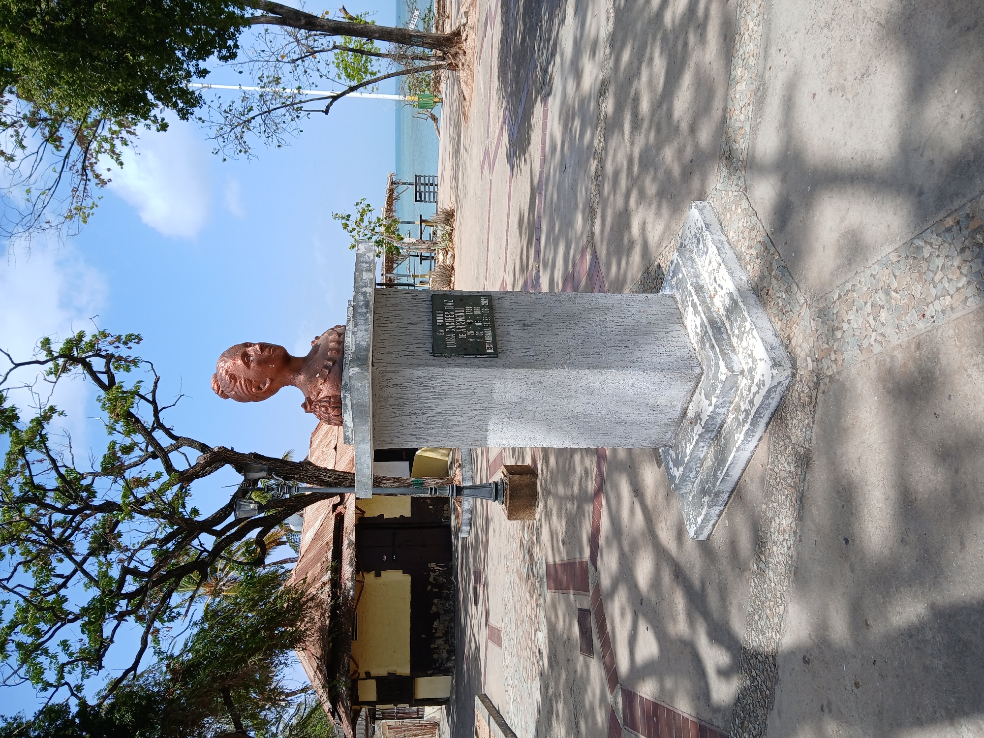The image depicts a copper bust of a woman on a concrete pedestal with a plaque, standing on a tiled walkway. The bust is a memorial to Luisa Cáceres de Arismendi, as indicated by the plaque, located in Juan Griego, Venezuela. The bust faces to the left of the frame. Behind the pedestal, there is a building with a weathered, corrugated metal roof and a partially visible storefront. A tree with bare branches is visible to the left. In the background, the ocean and coastline can be seen under a partially cloudy sky. Shadows from trees are cast on the walkway.