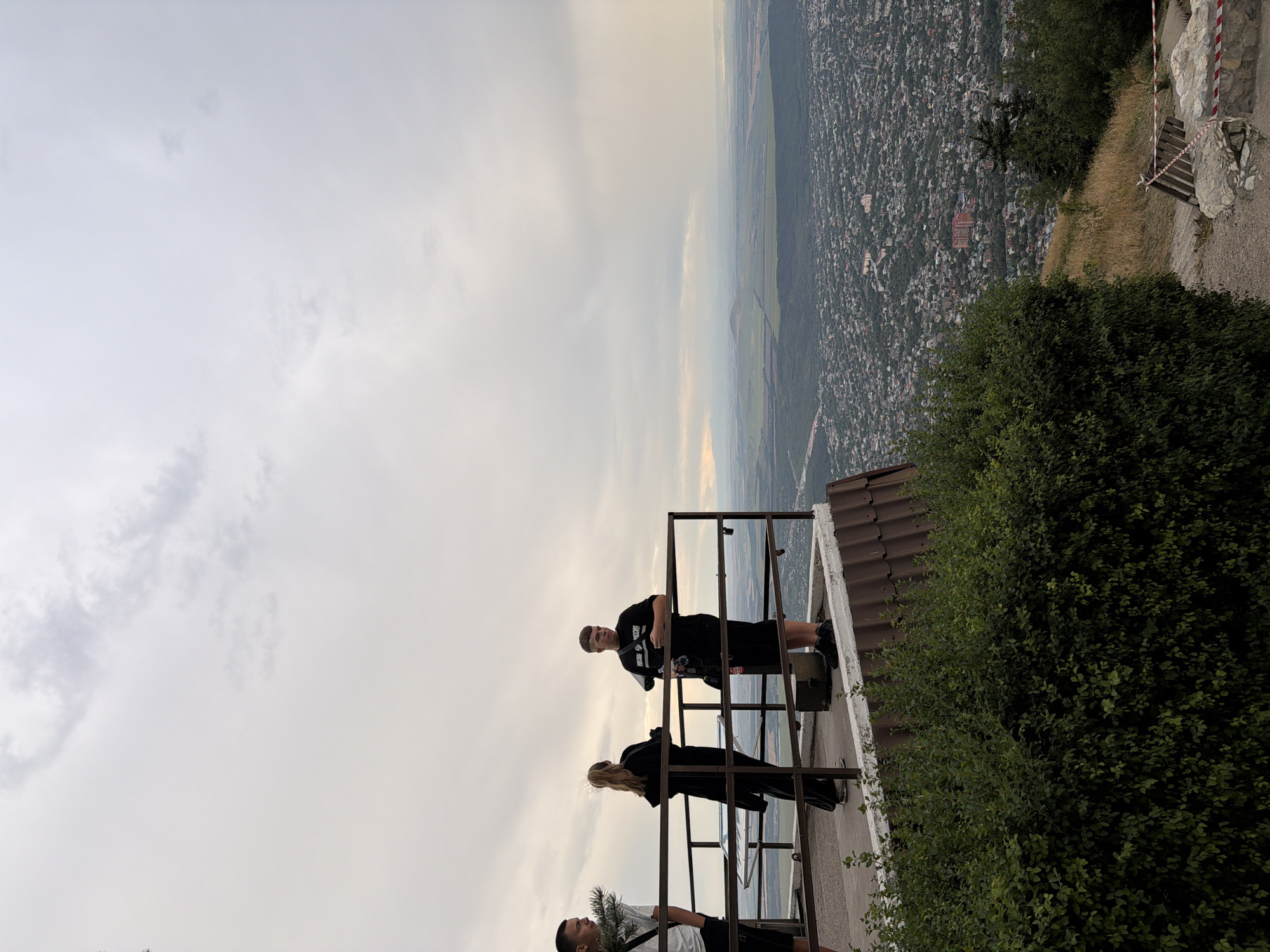Three individuals, two male and one female, stand on an elevated observation deck overlooking a city. The individuals are positioned near a metal railing. The male on the far left is mostly obscured by a plant. The female in the center is facing away from the camera, and the male on the right is facing towards the viewer, holding a camera. The observation deck has a tiled roof and is surrounded by lush green foliage. The city spreads out below the deck, with buildings and trees visible. In the distance, a vast landscape unfolds under a cloudy sky. The scene appears to be in Goryachevodsky, Russia, given the context provided by the original prompt.