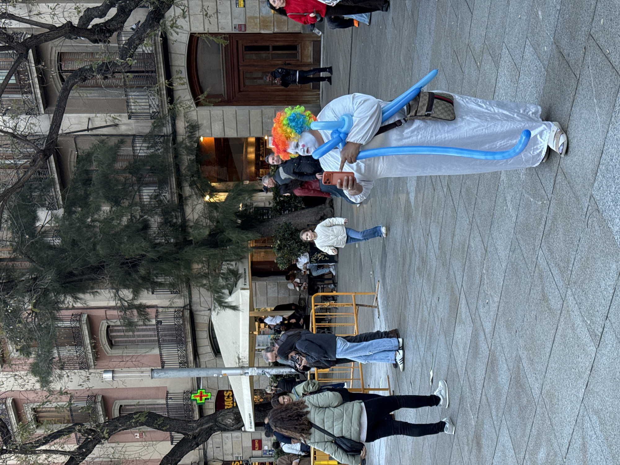 A street scene in Barcelona, Spain, shows a performer dressed as a clown wearing a rainbow wig, white face paint, and a white robe. The clown holds a smartphone in their right hand and two long blue balloons are wrapped around the neck and upper chest. A brown crossbody bag is also visible. Pedestrians walk by, some looking toward the clown, who stands on a paved sidewalk in front of a restaurant with outdoor seating, indicated by the yellow barriers. The buildings behind are typical of Barcelona architecture, featuring balconies and shutters. A pharmacy sign is visible on the left side of the frame. The street appears to be busy with pedestrian traffic.