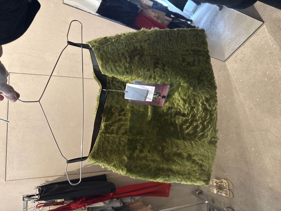 A Caucasian hand with short fingernails holds a lime-green, deep pile faux-fur mini skirt aloft on a silver wire hanger within a retail environment in Paris, France. The skirt features a dark brown internal waistband from which hang multiple tags via a silver chain, one clearly displaying "PRADA" and another featuring an image of a model wearing a similar green furry garment. To the left of the skirt, a metal clothing rack holds various garments, including a vibrant red dress and a black top. Beneath this rack, on the light grey concrete floor, a pair of metallic gold open-toe high-heeled sandals are visible. To the right, a tall mirror reflects parts of the store, including further garments (red and dark colors) and the lower legs and dark footwear of the person holding the skirt. The wall in the background is a light, untextured beige color.
