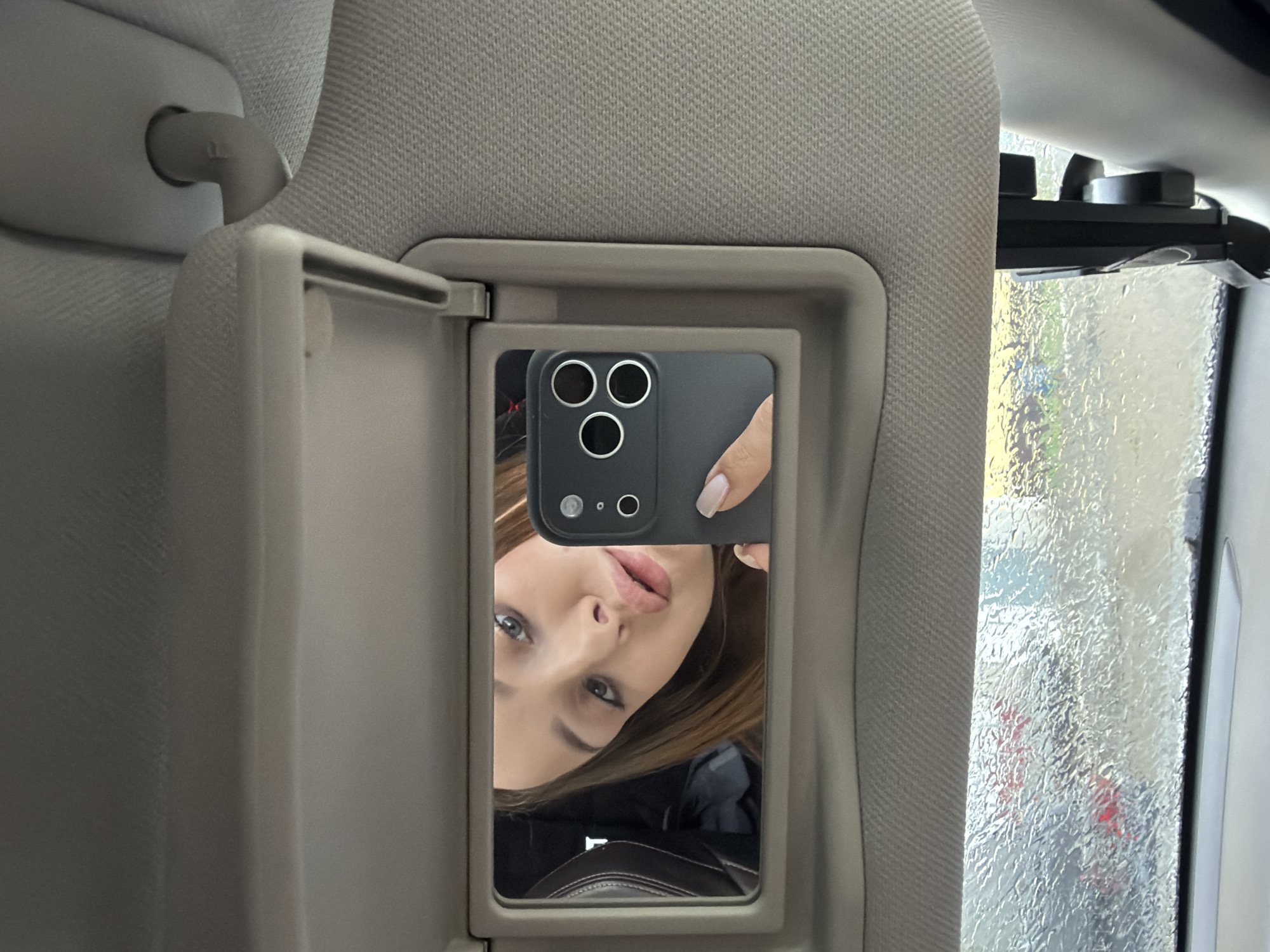 A selfie reflection in a car visor mirror shows a woman with fair skin, light brown hair, and grey eyes, holding a black smartphone with three camera lenses. The visor and surrounding car interior are beige. Outside the car window, rain is visible, obscuring the view. It's not possible to determine the location is Chaikovsky, Russia from this image alone.