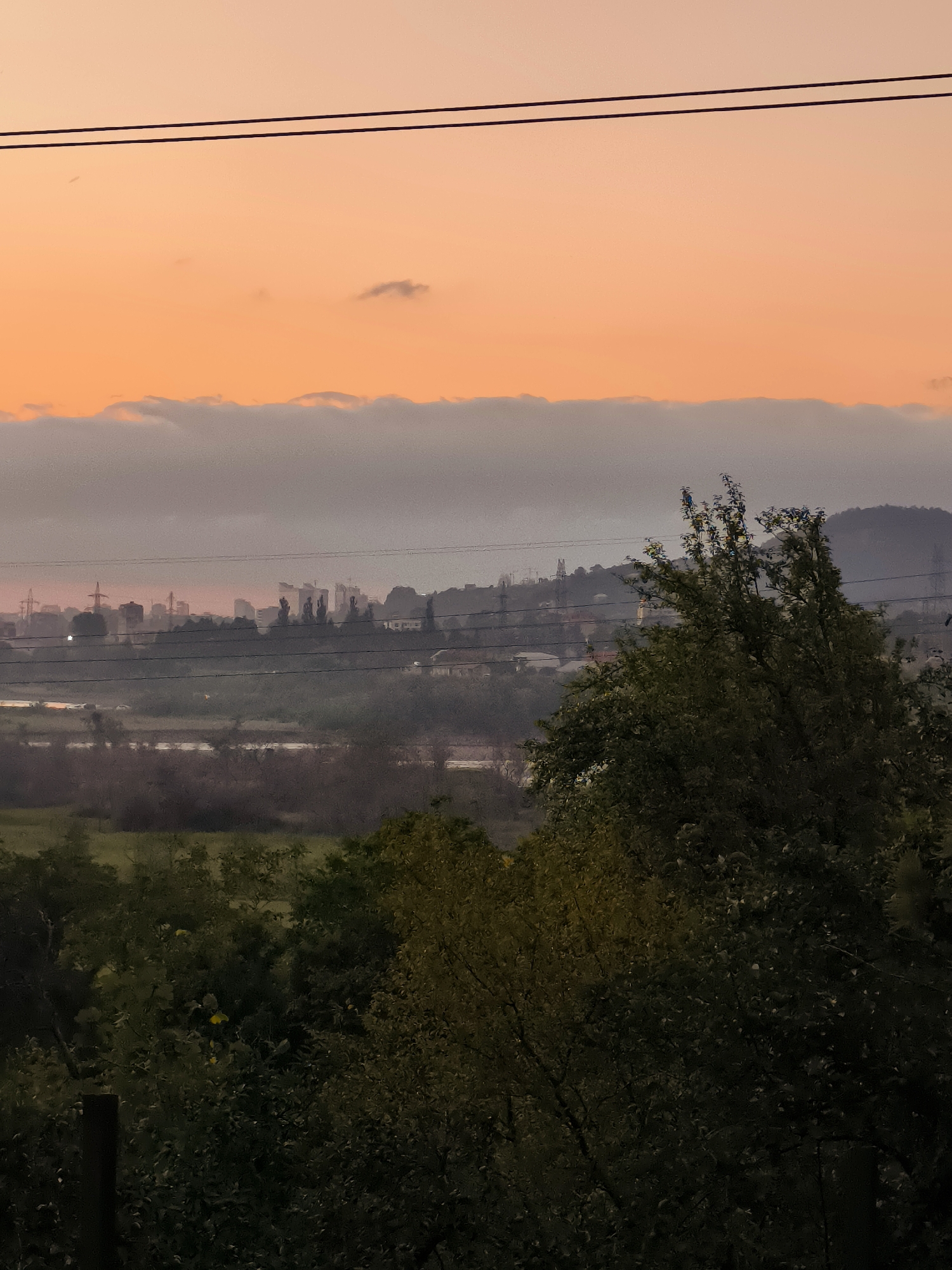 A rural landscape is depicted at dusk, featuring a prominent orange sky with a band of low-lying clouds separating it from the hazy cityscape in the distance. Two thin, dark power lines traverse the upper portion of the image. In the foreground, lush green trees obscure much of the lower half of the frame, with a dark wooden post visible on the far left. The middle ground shows a body of water and more vegetation, leading towards the distant urban area. The cityscape itself is indistinct, with silhouettes of buildings and industrial structures visible through the atmospheric haze. The overall impression is one of a serene, yet industrial, twilight scene in Makho, Georgia.