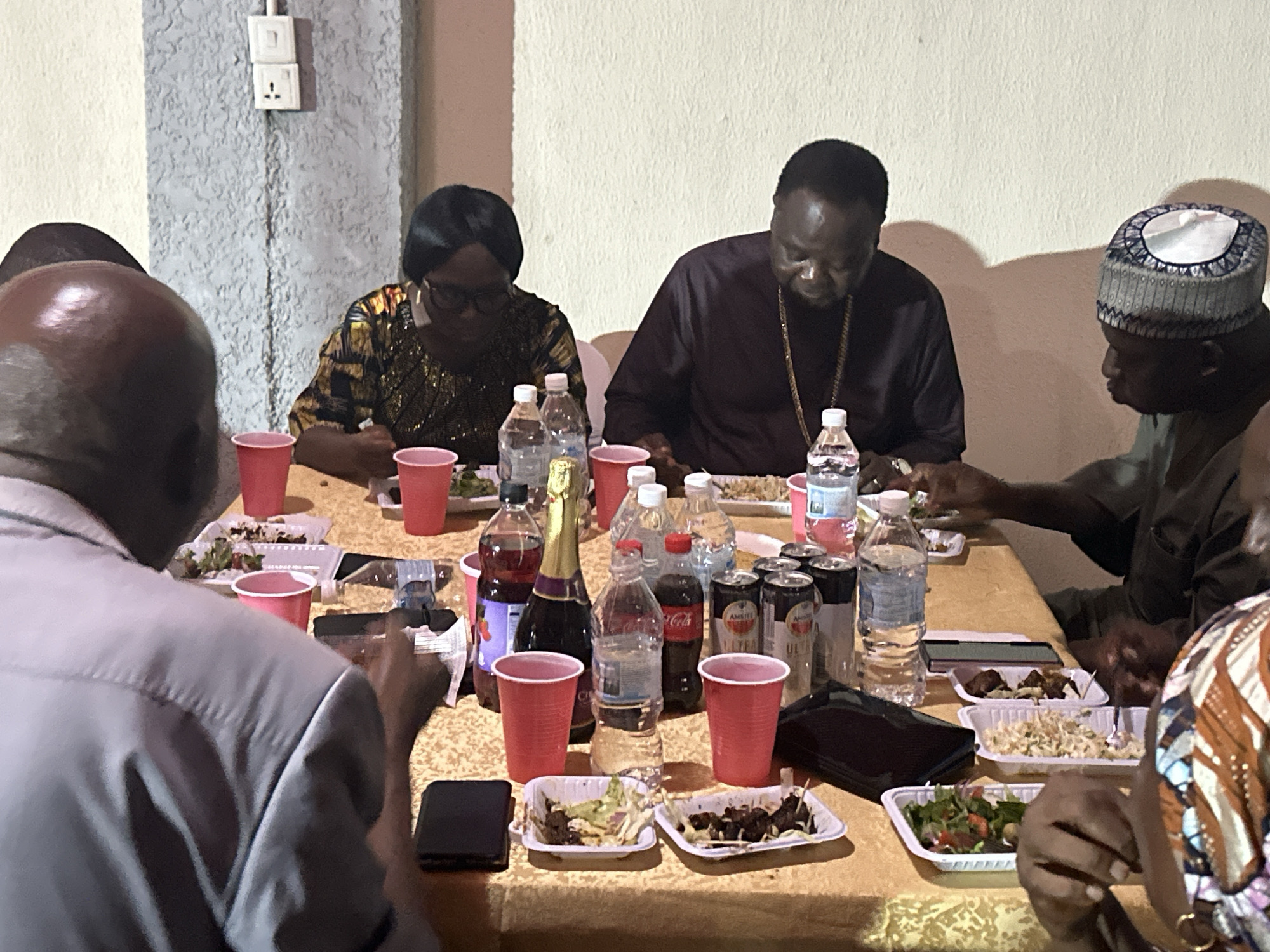 A group of people are sitting around a table in a dimly lit room in Narayi, Nigeria. The table is covered with a gold-colored tablecloth and is laden with food and drinks, including water bottles, soda cans, juice, and food served in disposable white trays. Red plastic cups are scattered across the table. The individuals around the table appear to be eating. One person wears a traditional cap, another has a gold necklace, and a woman wears glasses. A power outlet is visible on the wall in the background.