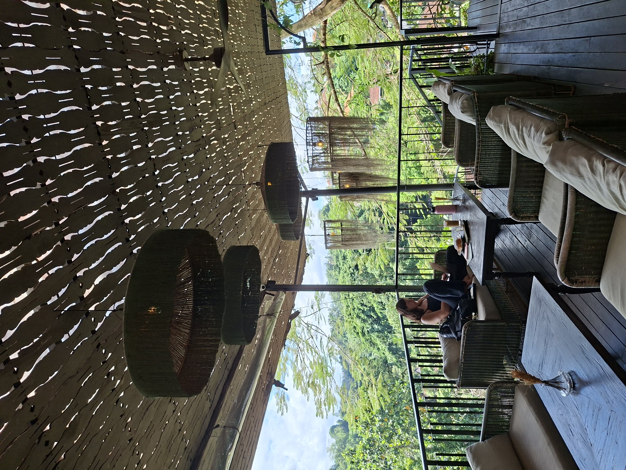 A woman sits on a rattan chair at a table on a wooden deck, overlooking lush greenery, likely in Ubud District, Indonesia. The deck, featuring rattan furniture with cushions and a black iron railing, is partially covered by a ceiling with long horizontal slits. Hanging from the ceiling are several circular woven light fixtures. Additional decor includes glass vases on the tables. The environment outside the railing shows dense foliage.