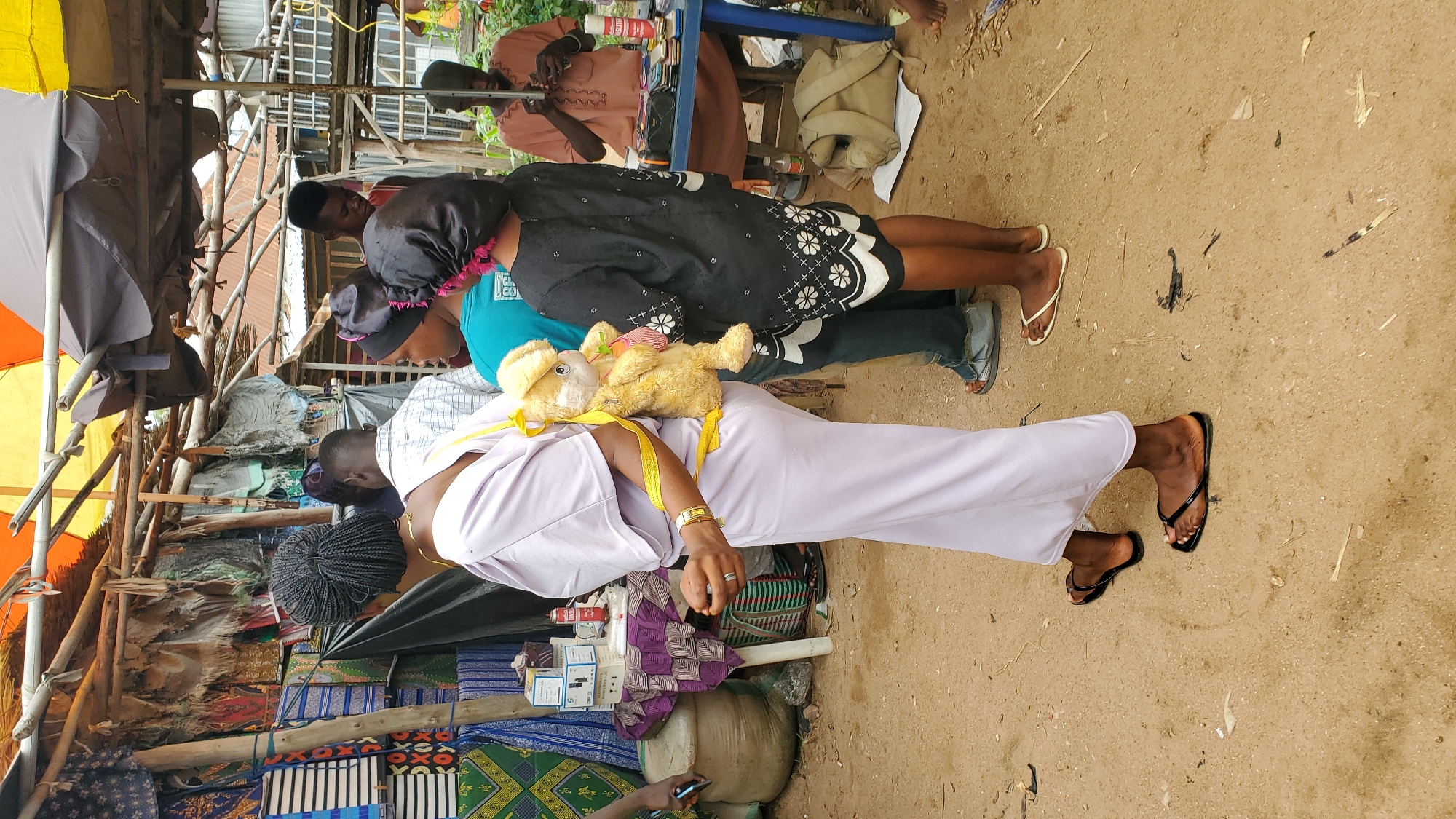 In Jalingo, Nigeria, this image depicts a bustling outdoor market scene under a makeshift canopy constructed from wood and tarpaulin. The ground is covered in dry, sandy earth. In the foreground, a woman wearing a pale purple dress and black flip-flops carries a stuffed yellow bear on her back, secured with a yellow strap. To her right, a young person in a black dress with floral embroidery and a black headscarf stands with their back to the camera. Behind them, other individuals are visible, including a person in a blue shirt and another in a plaid shirt. Further back, a man in a brown garment sits at a blue table laden with various items, possibly for sale. The market stalls display textiles and other goods, with stacks of fabric and packaged items visible. The overall impression is one of everyday commerce and community interaction in an informal setting.