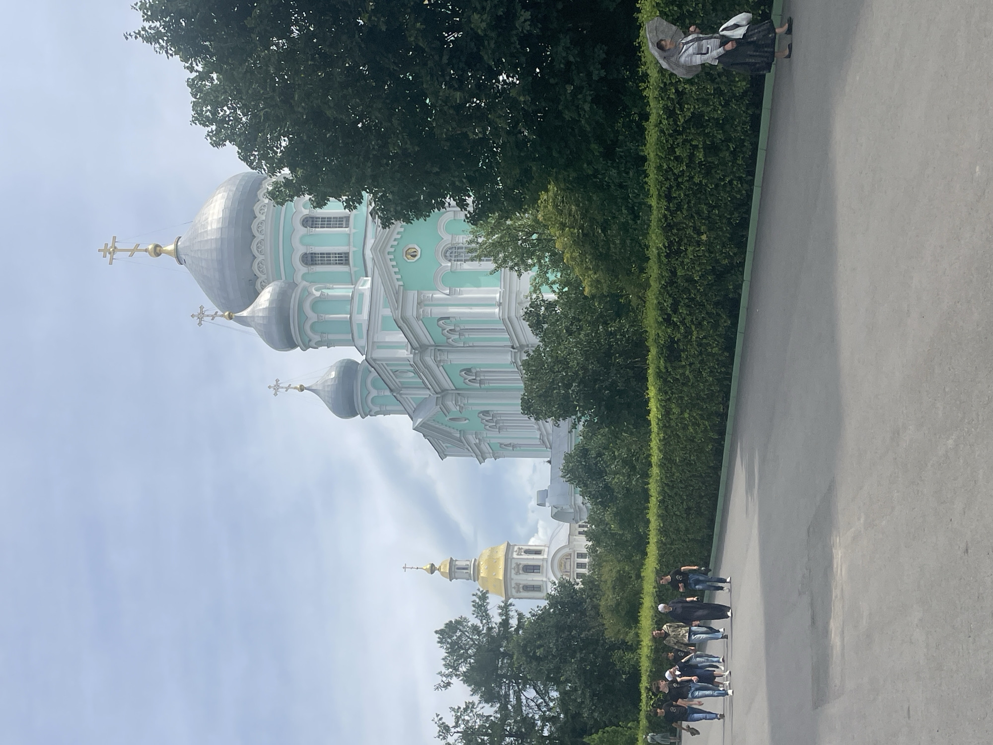The image shows a group of people walking along a paved path in front of a large, ornate church with multiple domes in Diveyevo, Russia. The church is predominantly light teal and white, with silver and gold domes. A taller, separate bell tower with a golden dome is visible to the left of the main church. Lush green trees and hedges line the path. In the foreground, a group of young people, appearing to be teenagers and young adults, walk together, some looking at their phones, others talking. Further down the path, a woman walks alone, holding an umbrella. The sky is overcast, with scattered clouds.