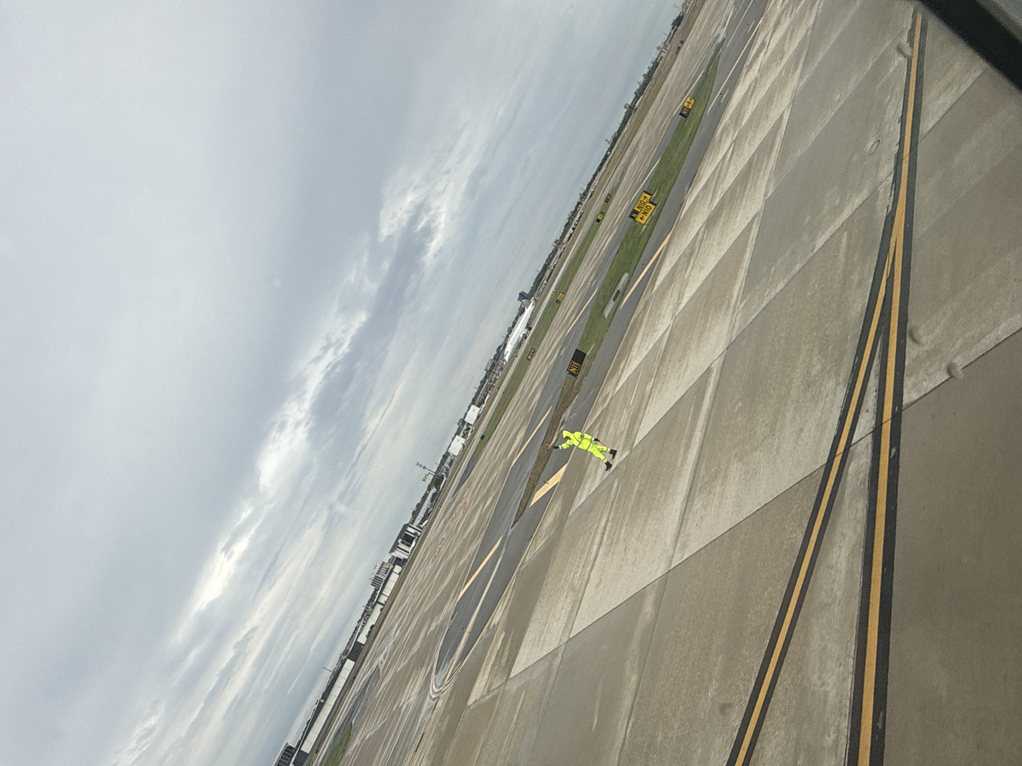 The image depicts a view from an elevated position, likely an aircraft window, overlooking an airport tarmac under an overcast sky. The tarmac is concrete and marked with yellow and black lines. Small patches of grass are visible between the concrete sections. Yellow signs with black lettering are interspersed throughout the tarmac. A person wearing a high-visibility yellow suit is visible on the tarmac. The general location is the airport in San Antonio, United States of America.
