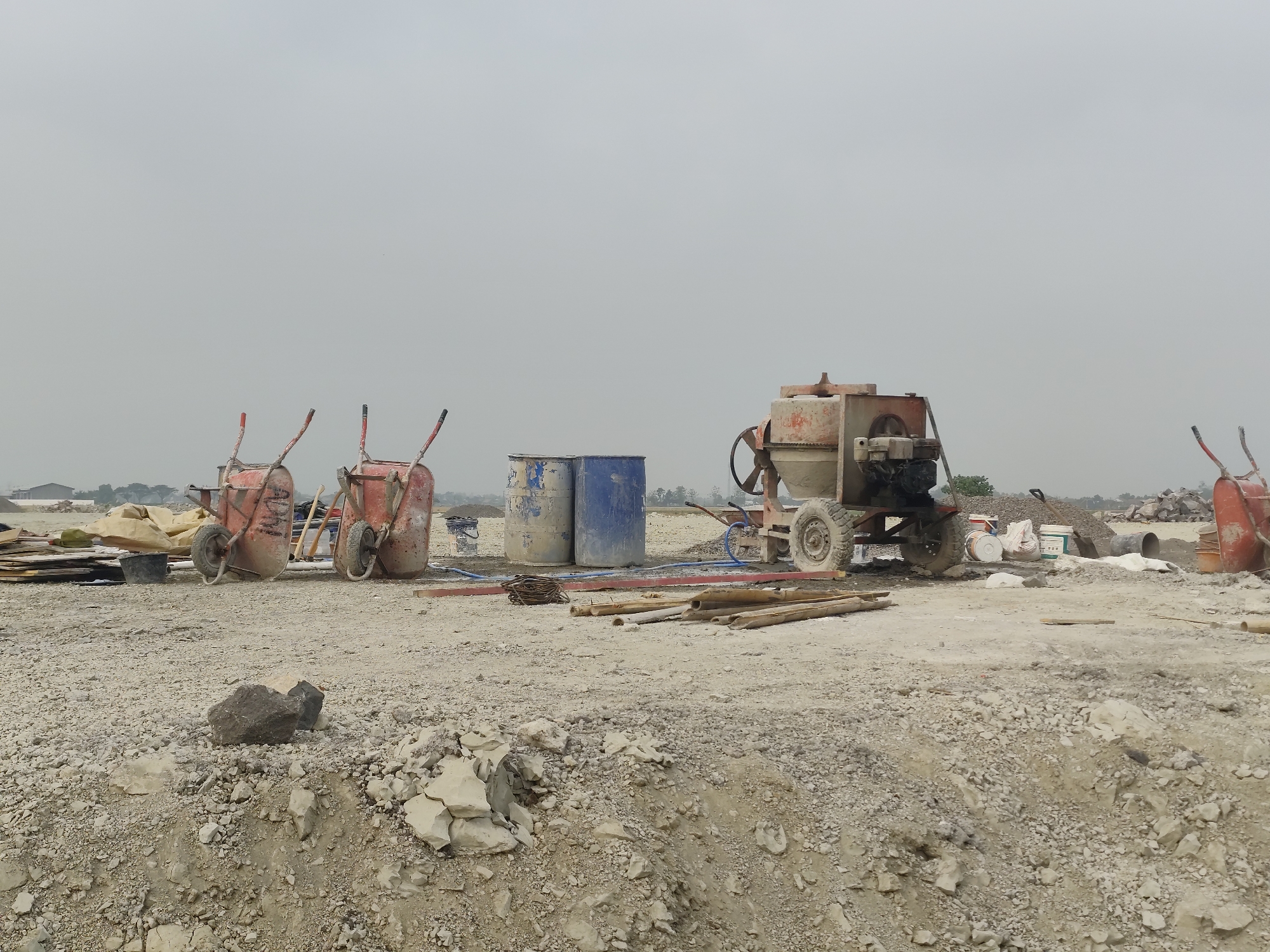 The image depicts a construction site in Purwodadi, Indonesia, under an overcast sky. The ground is covered with gravel and dirt, with piles of debris and construction materials scattered throughout. In the foreground, a large rock rests on the gravel. In the mid-ground, two red wheelbarrows with metal basins and handles are visible, one positioned slightly behind the other. Further to the right, two cylindrical blue and white barrels stand side-by-side, with a red and white cement mixer to their right. The cement mixer has a rotating drum and is mounted on a wheeled chassis with a visible engine. Several wooden planks and pipes are also present on the ground. In the background, more construction equipment and materials can be seen, along with a few structures and trees in the distance. The overall scene is one of active construction, with an absence of people.