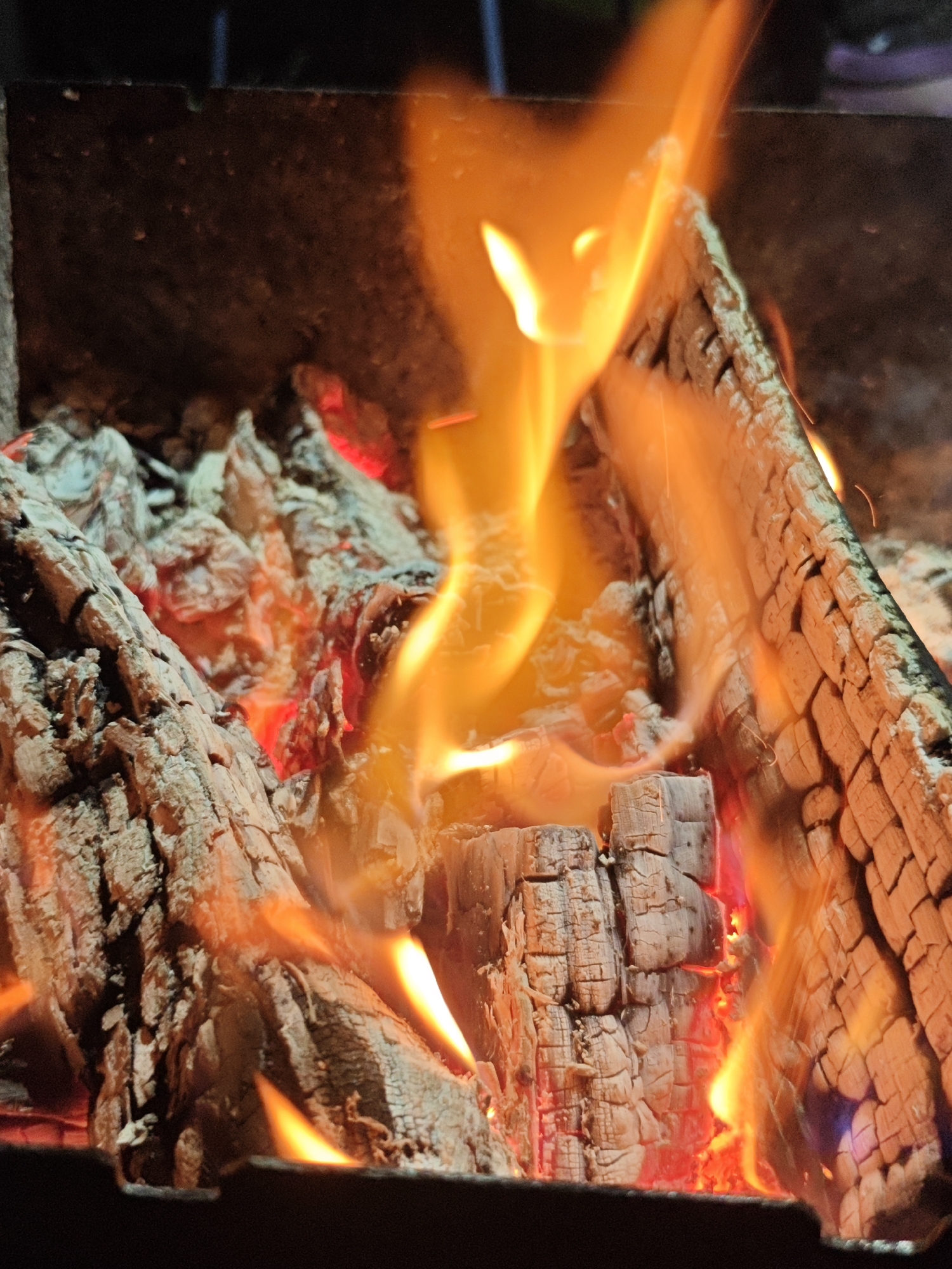 A close-up, low-angle, indoor shot depicts logs burning in a fire pit. The logs are charred and cracking, with embers glowing red beneath. Bright orange flames lick upwards, obscuring some of the wood and casting a warm glow on the surrounding area. The background is dark and out of focus. The scene is likely in a domestic setting given the size and style of the fire pit. There are no people present.