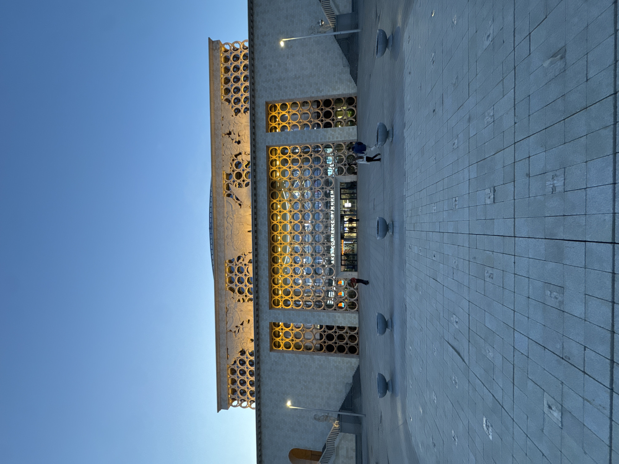 The image shows the facade of the State Central Concert Hall "Russia" in Moscow, Russia, taken from a low angle. The building features a grid of circular openings, with golden light emanating from within. A small group of people is visible near the entrance. The exterior is constructed from light-colored stone or concrete. A paved plaza extends in front of the building. The sky is clear and light blue.
