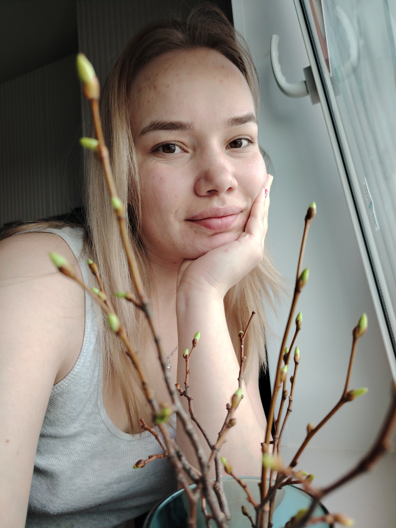A woman with light brown hair, light skin, and freckles poses for a selfie indoors near a window. She rests her chin on her right hand, her expression calm and slightly smiling. A vase filled with budding branches is positioned in the foreground, partially obscuring her. The background includes a window with a white frame and a wall with vertical lines. There is no discernible evidence within the image to confirm the location is Марковский, Russia.
