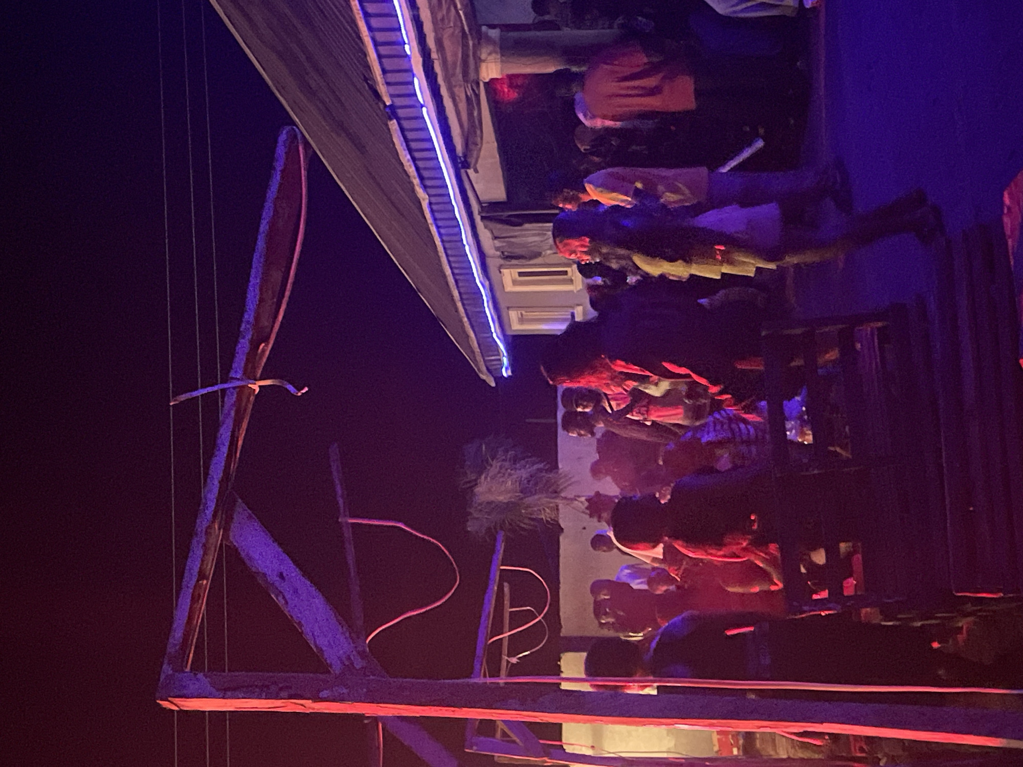 The image captures a nighttime gathering of people in Durumi, Nigeria. A wooden structure, partially illuminated by red and purple lights, stands prominently in the foreground, with a rope hanging from it. In the background, a building with a corrugated metal roof is visible, adorned with blue neon lights along its eaves. A crowd of individuals is dispersed around the area, some standing and others seated on wooden benches. The atmosphere appears to be social and relaxed, with people engaging in conversation and general assembly. The lighting conditions suggest an outdoor event or celebration.