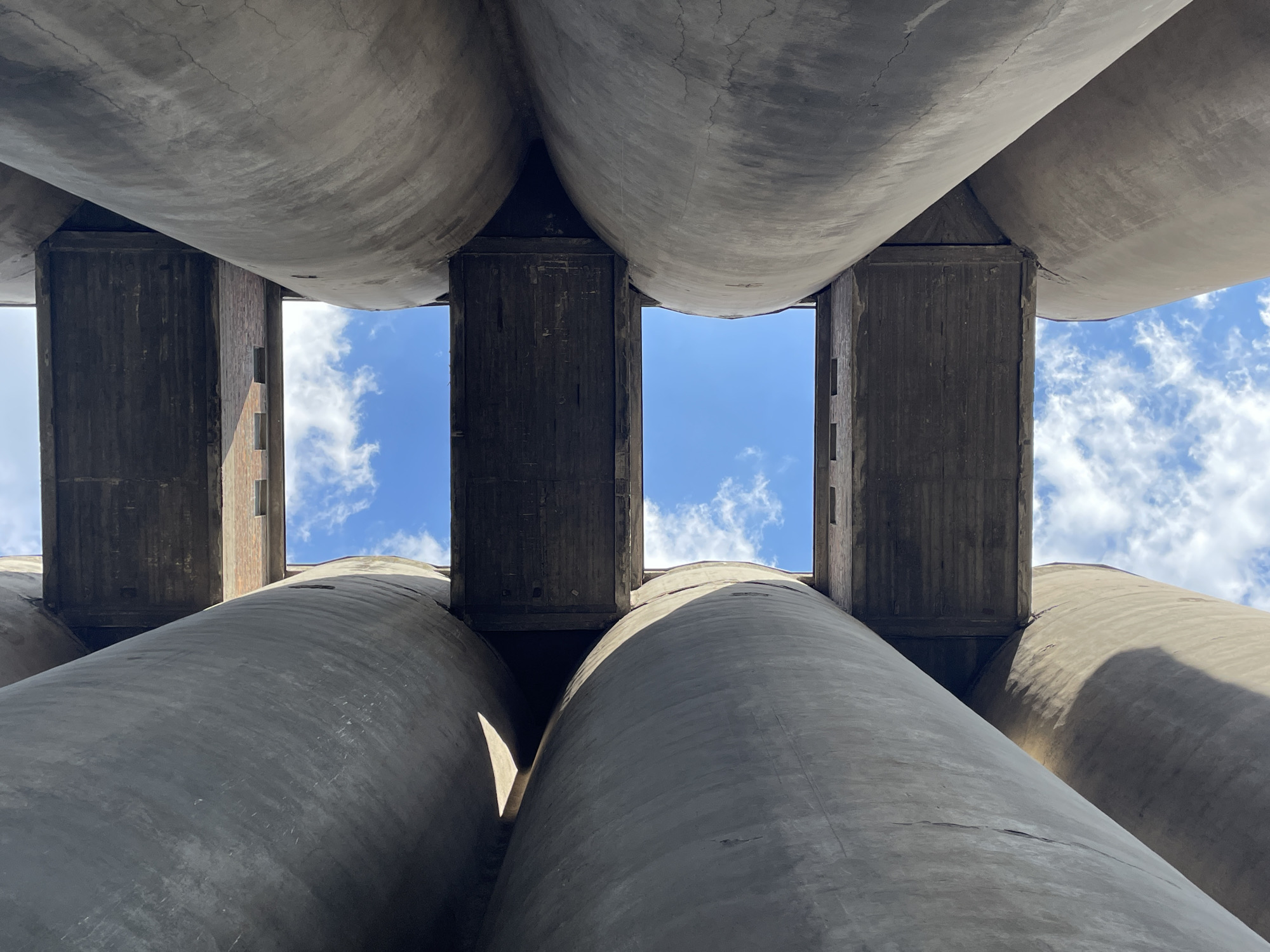 The image is an upward-looking view of the concrete silos at the Museum of Contemporary Art in Belgrade, Serbia. The perspective creates a narrowing effect, with cylindrical concrete structures dominating the frame. Rectangular concrete bridges or platforms are interspersed between the silos, forming openings through which patches of blue sky with scattered white clouds are visible. The concrete is cracked and weathered, indicating age and exposure to the elements. There are no people visible.