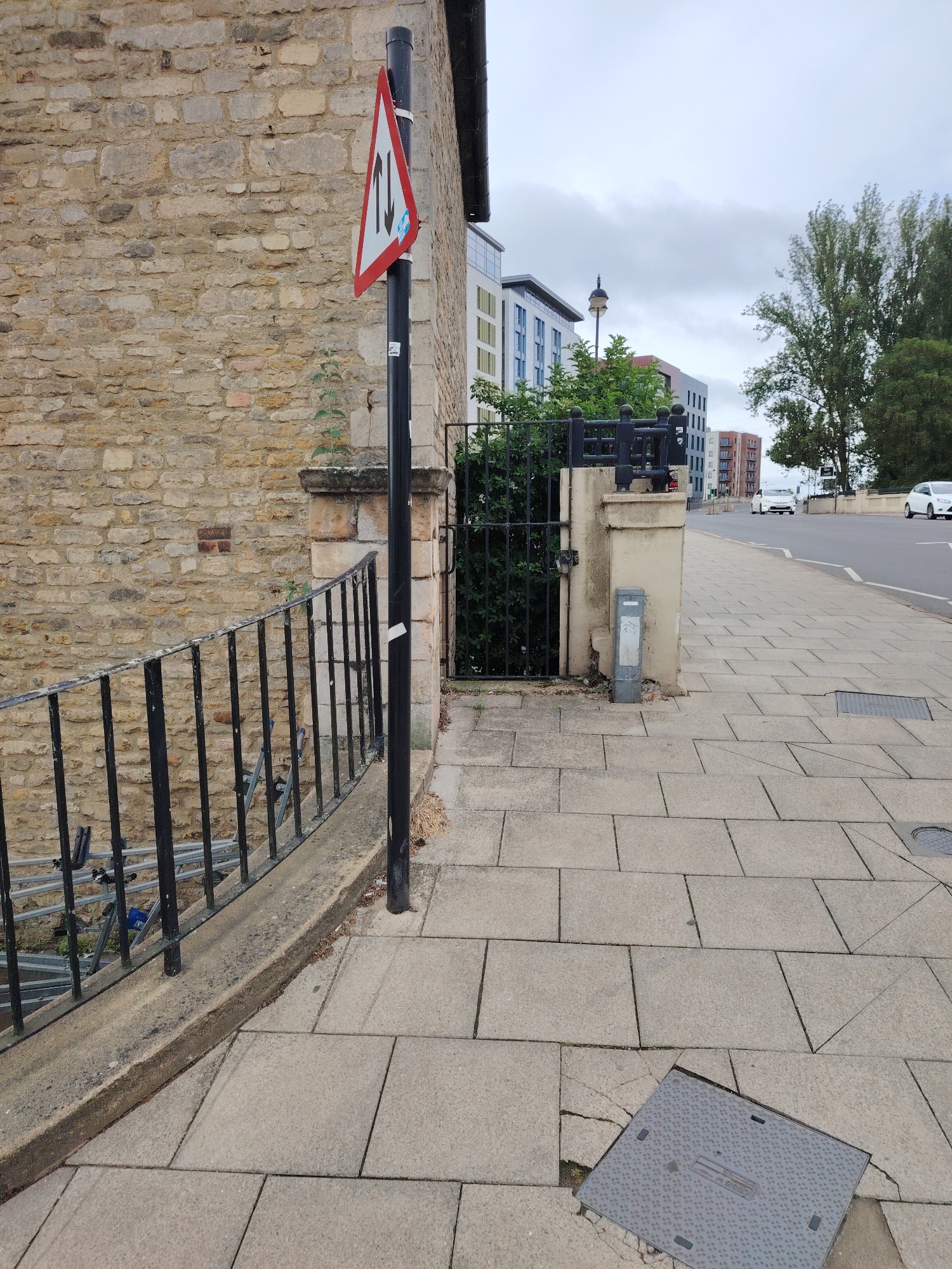 This image presents an outdoor urban scene under an overcast sky, located in Peterborough, United Kingdom. In the immediate foreground on the left, a black metal railing, consisting of vertical bars, runs along a low concrete wall that shows signs of weathering and minor discoloration. Beneath the railing, metallic scaffolding components are partially visible. A black, cylindrical pole stands upright in the mid-ground, anchored to the ground. A triangular road sign is affixed to this pole, featuring a red border, a white interior, and two vertical black arrows—one pointing upwards and the other downwards—indicating a two-way traffic condition.
Adjacent to the pole and extending to the left, a substantial wall constructed from irregularly shaped, light brown and grey stone blocks forms part of an older structure. A small green plant sprouts from a crevice in this stone wall. A dark gutter system is visible along the upper edge of the building to the far left.
The central and right portions of the image are dominated by a paved sidewalk made of rectangular grey slabs, extending into the background. Some slabs exhibit minor cracks and unevenness. A square, dark grey metal utility access cover, embossed with a textured pattern and small rectangular cutouts, is embedded in the sidewalk in the bottom right foreground, surrounded by fractured pavement.
Further along the sidewalk, a concrete pillar supports a black metal gate, which is largely obscured by dense green foliage behind it. A grey, cylindrical metal bollard is attached to the base of the concrete pillar. Beyond the gate and vegetation, a modern, multi-story building with a light-colored facade (incorporating white, light blue, and possibly muted green sections) and multiple windows is visible. A street lamp is situated between this building and others in the distant background.
A paved road runs parallel to the sidewalk on the right, delineated by a white edge line. Two white passenger vehicles are present on the road: one in the far distance proceeding away from the viewer, and another closer in the mid-ground. In the background, a cluster of tall, leafy green trees stands to the right of the road, their foliage appearing to be in motion. Additional multi-story buildings are faintly visible in the far distance beyond the trees.