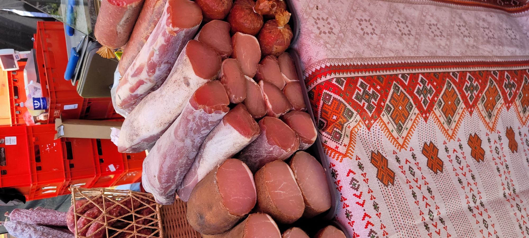 The image depicts a market stall overflowing with various cured meats, likely at the Riga Central Market given the red crates visible in the background which are characteristic of the market's layout. In the foreground, a variety of cured meat products are displayed on a table covered with a patterned tablecloth. The meats are arranged in stacks, with some whole cured sausages in a woven basket on the left. The cuts of meat range from thick, rod-shaped items with a white exterior and pink interior to smaller, sliced pieces of cured pork. The background shows multiple stacks of red plastic crates, a scale, and what appears to be a bottle of liquid. The lighting suggests an outdoor or partially covered setting.