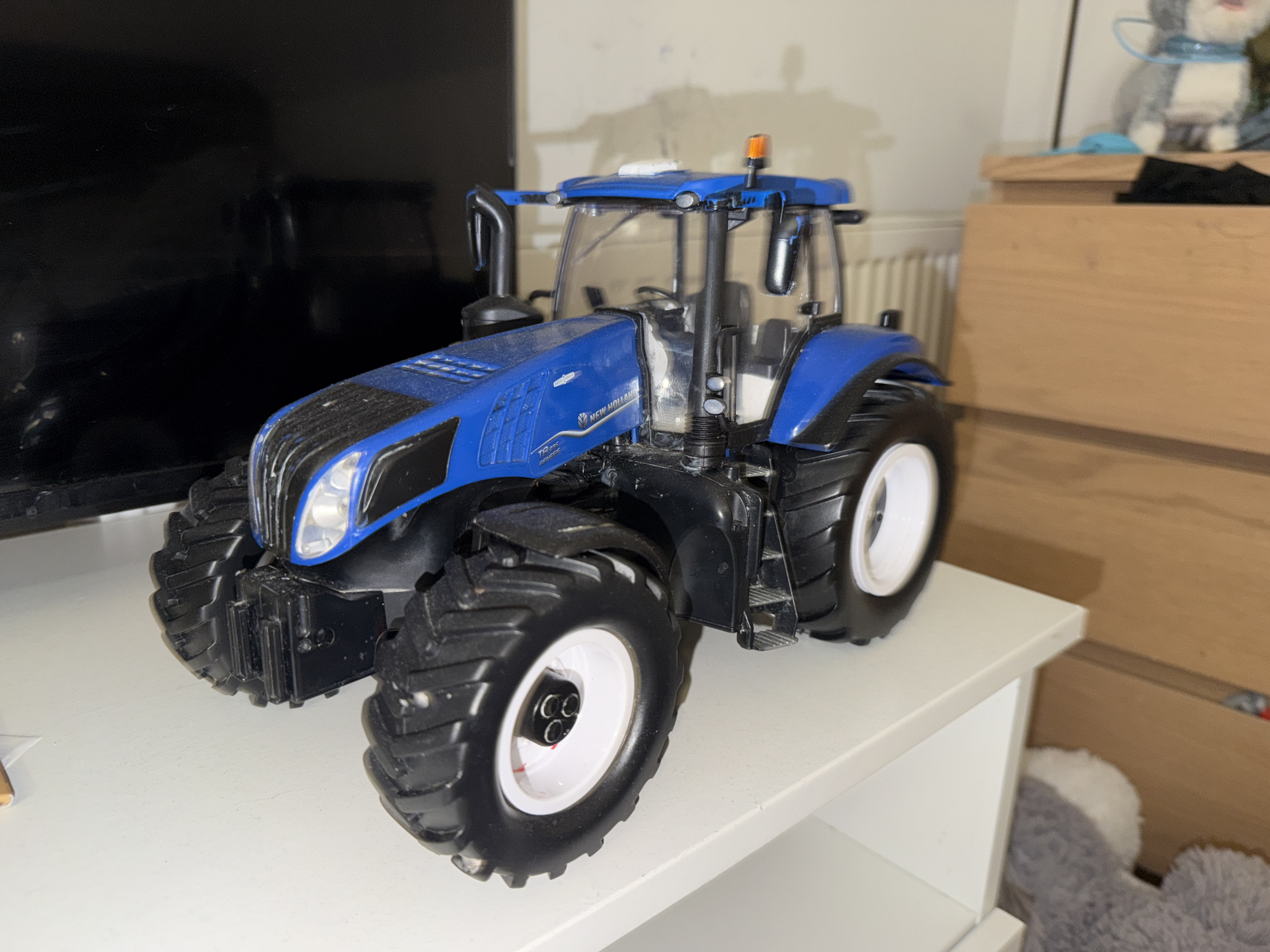 A blue and black toy tractor is positioned on a white shelf. The tractor is detailed, with visible tires, cabin, and engine parts. In the background, a dark surface, possibly a television screen, is to the left, and a wooden dresser is to the right. A portion of a white radiator is also visible behind the tractor. The scene appears to be indoors, likely in a residential setting.
